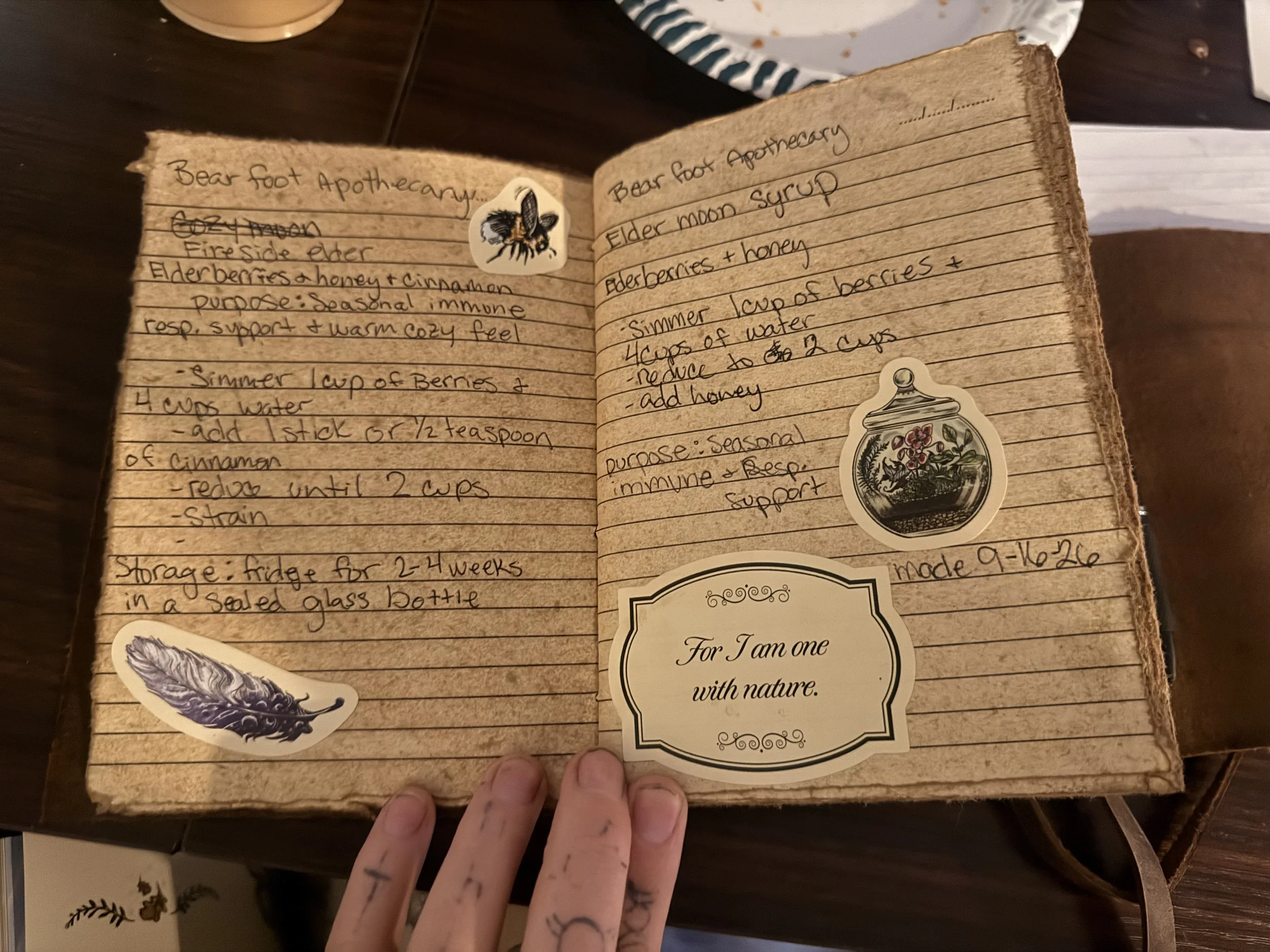 Open handwritten herbal recipe journal labeled “Bear Foot Apothecary,” featuring elderberry syrup recipes, vintage nature stickers, and a hand-drawn bee.