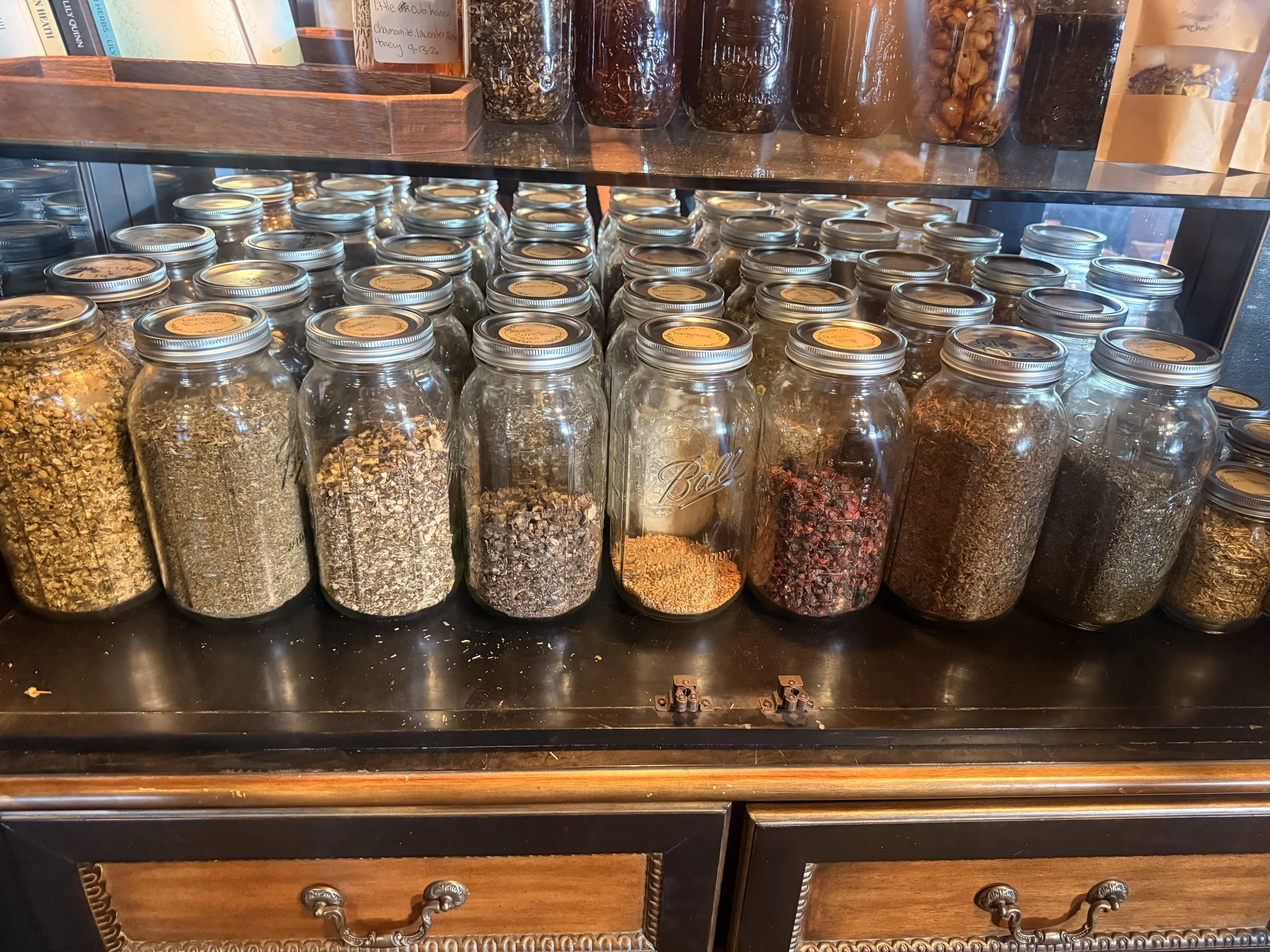 Rows of labeled glass jars filled with dried herbs, flowers, and spices in a wooden display cabinet