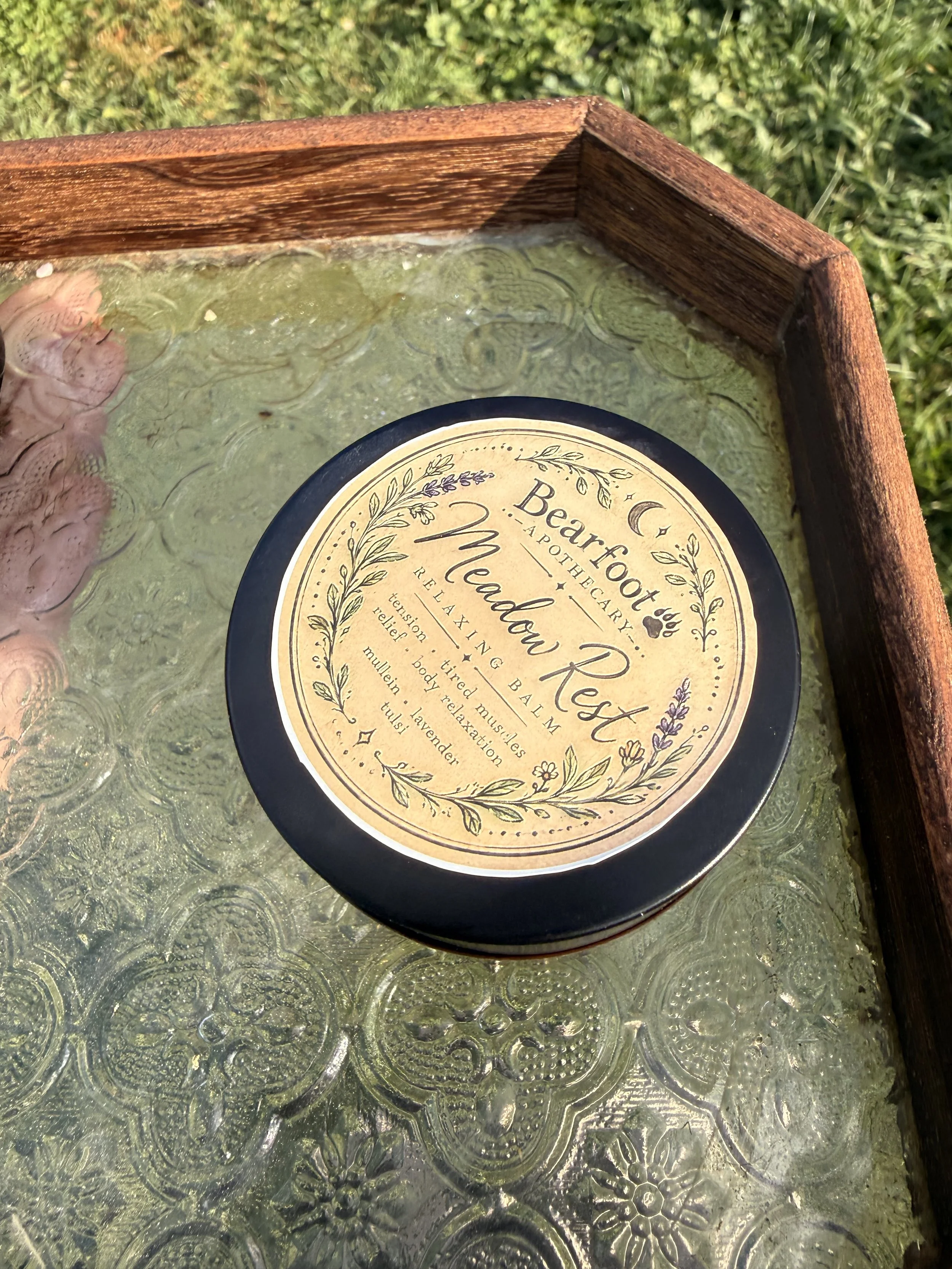 Close-up of a vintage wooden tray with a floral keepsake plaque on a textured glass tabletop, surrounded by greenery.