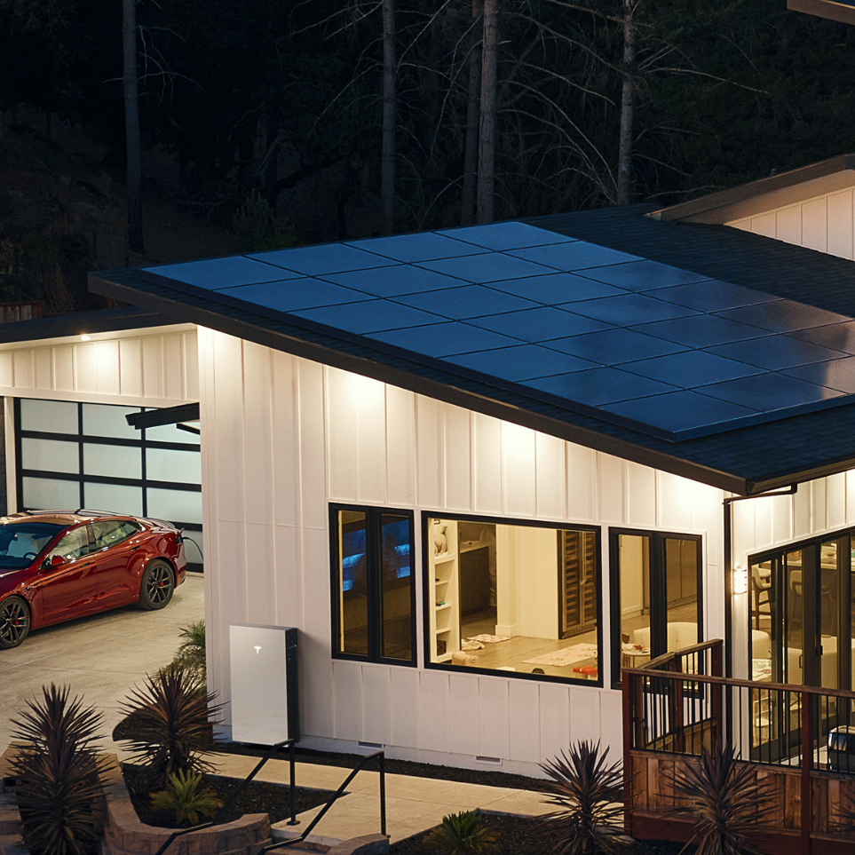 A modern house with white siding, large windows, and solar panels on the roof, with a red sports car in the garage and plants in the front yard at dusk.