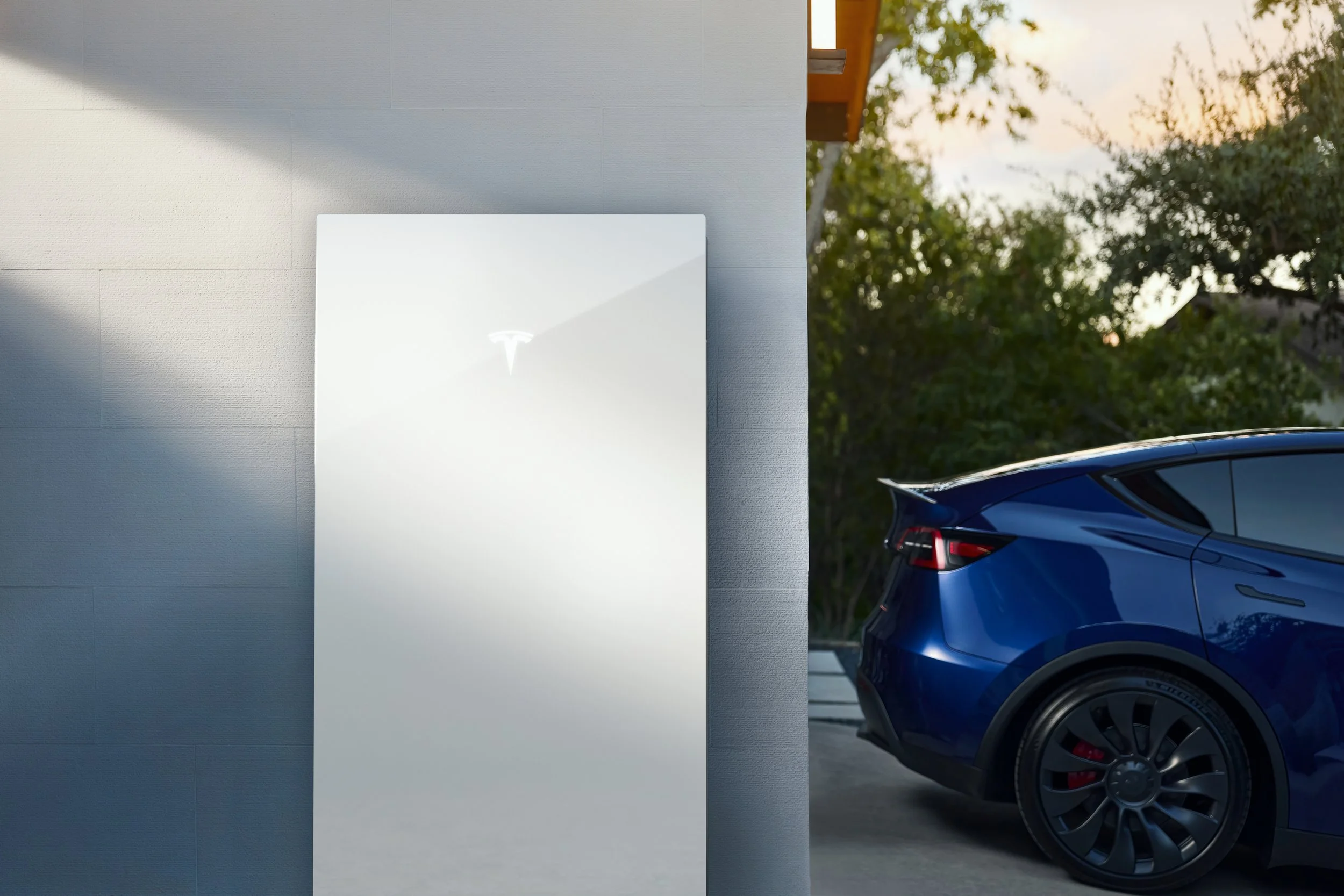 A modern white building with a Tesla vehicle parked nearby, and trees with green leaves in the background during sunset.
