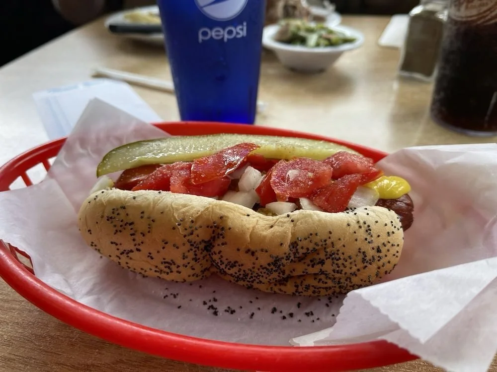 Hot dog in a poppy seed bun with ketchup, mustard, diced onions, relish, and pickles in a red basket with a Pepsi drink in the background.