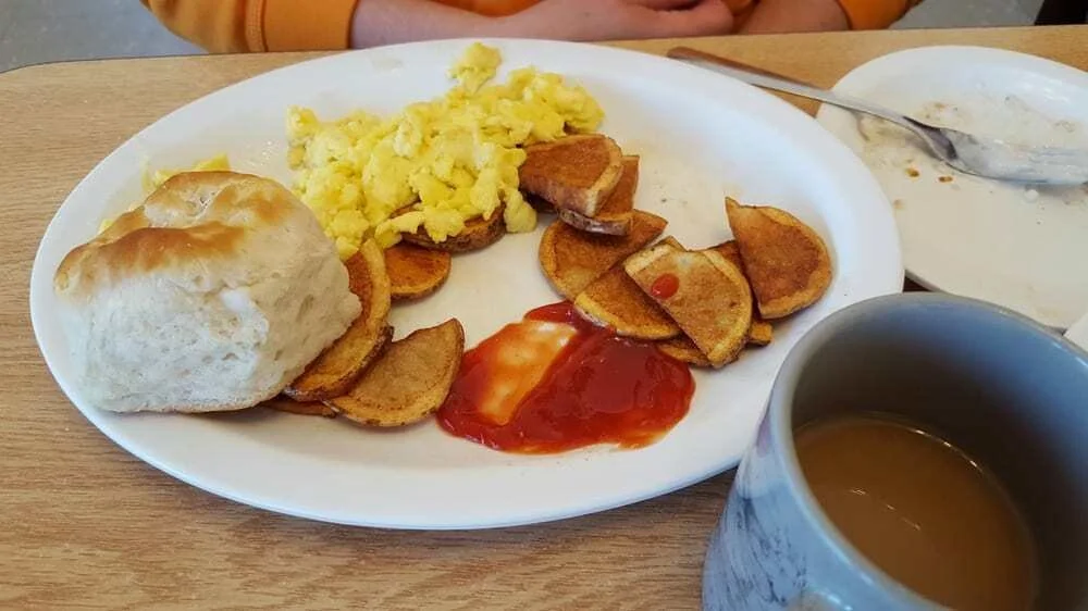 A breakfast plate with scrambled eggs, sliced potatoes, a bread roll, ketchup, and a side of empty or partially eaten bowl, alongside a mug of coffee on a wooden table.
