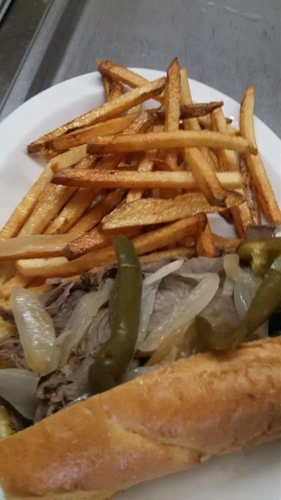 A plate of food with French fries, cooked onions, green pickles, and a partially visible bread roll or sandwich.