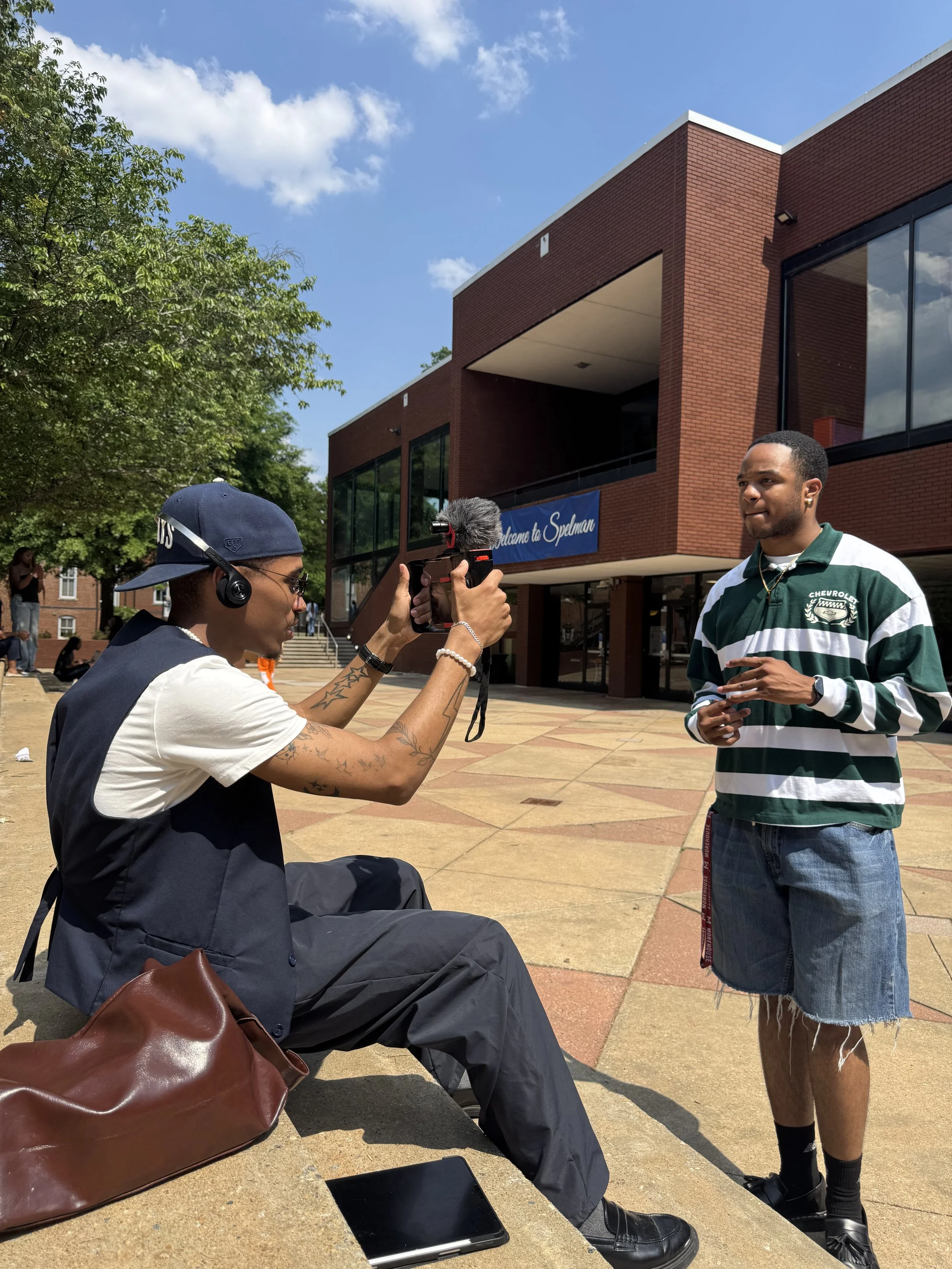 A young man with a striped green and white shirt is talking to a person sitting on a bench, who is filming him with a handheld camera. The person sitting is wearing a dark vest, glasses, a cap, and has tattoos on his arms. There are trees, a blue sky with clouds, and a brick building with a sign that reads "Welcome to Spelman" in the background.