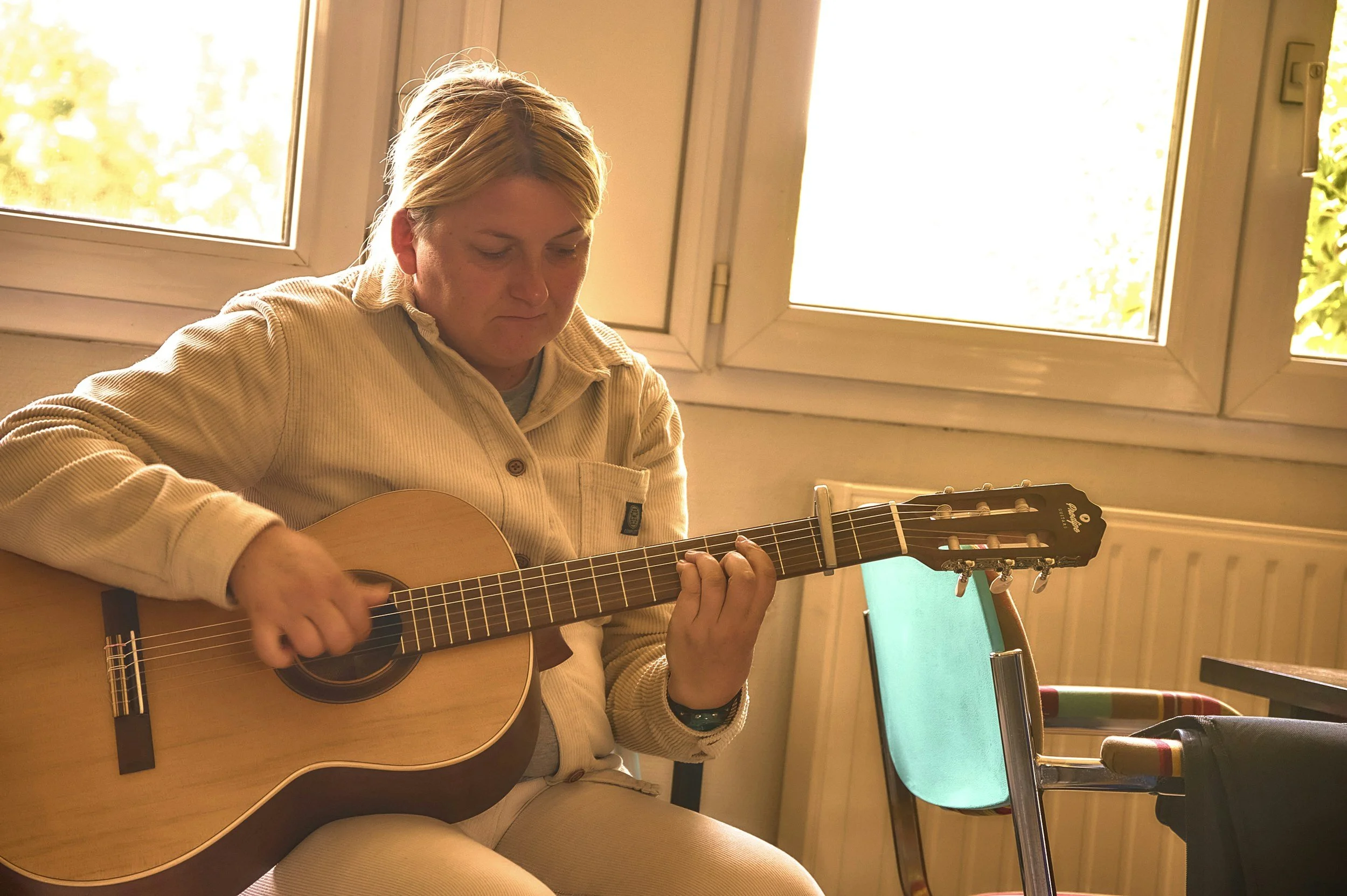 A woman wearing a beige jacket playing an acoustic guitar indoors near a window.