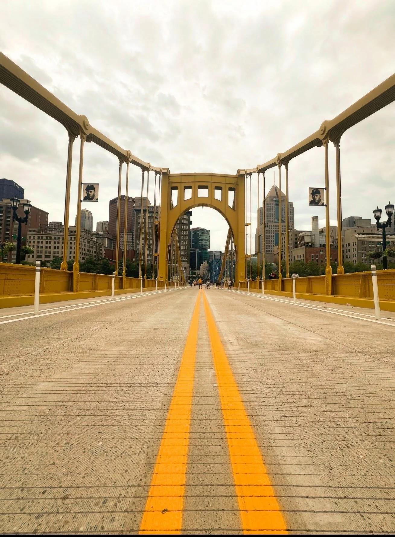 #3 Yellow bridge with suspension cables, city skyline in the background, cloudy sky, and a street with yellow lines.