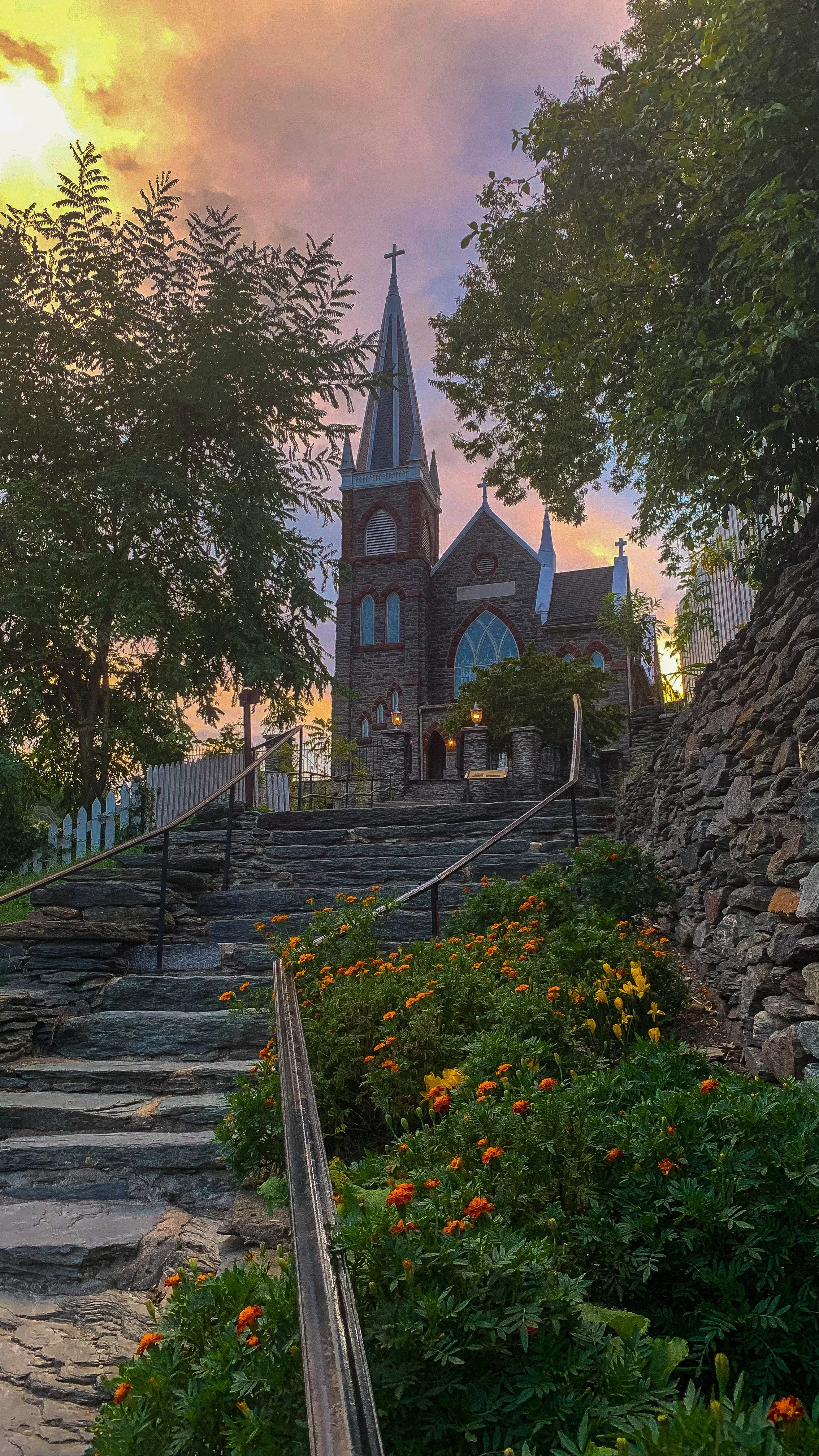#11 A stone church with tall steeples and crosses on top, viewed from the bottom of stone steps surrounded by orange flowers and green trees at sunset or sunrise.
