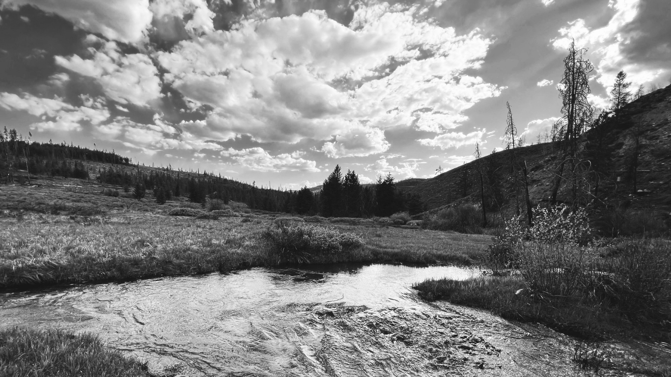 A black and white landscape photo of a small river flowing through a grassy meadow, with hills and trees in the background and a cloud-filled sky overhead.