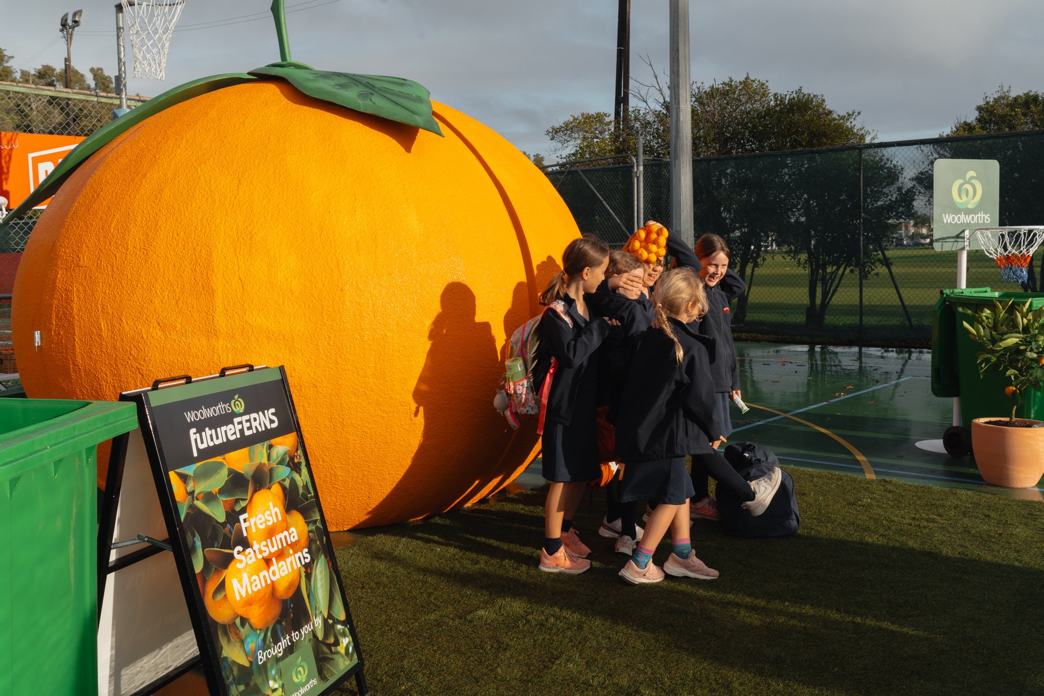 Group of schoolchildren standing beside a large mandarin structure on an outdoor sports court. Sign advertising 'Fresh Satsuma Mandarins' nearby, an activation by Dentsu for Woolworths New Zealand.