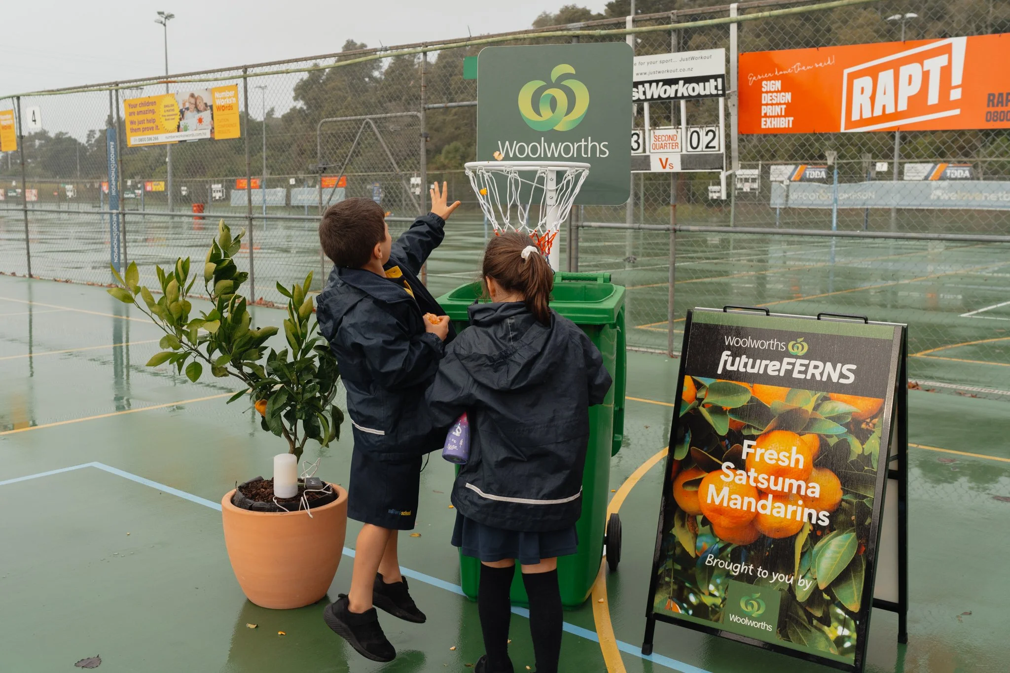 Two children in school uniform play basketball near a green trash bin on a wet outdoor sports court, with advertisements and a basketball hoop in the background.