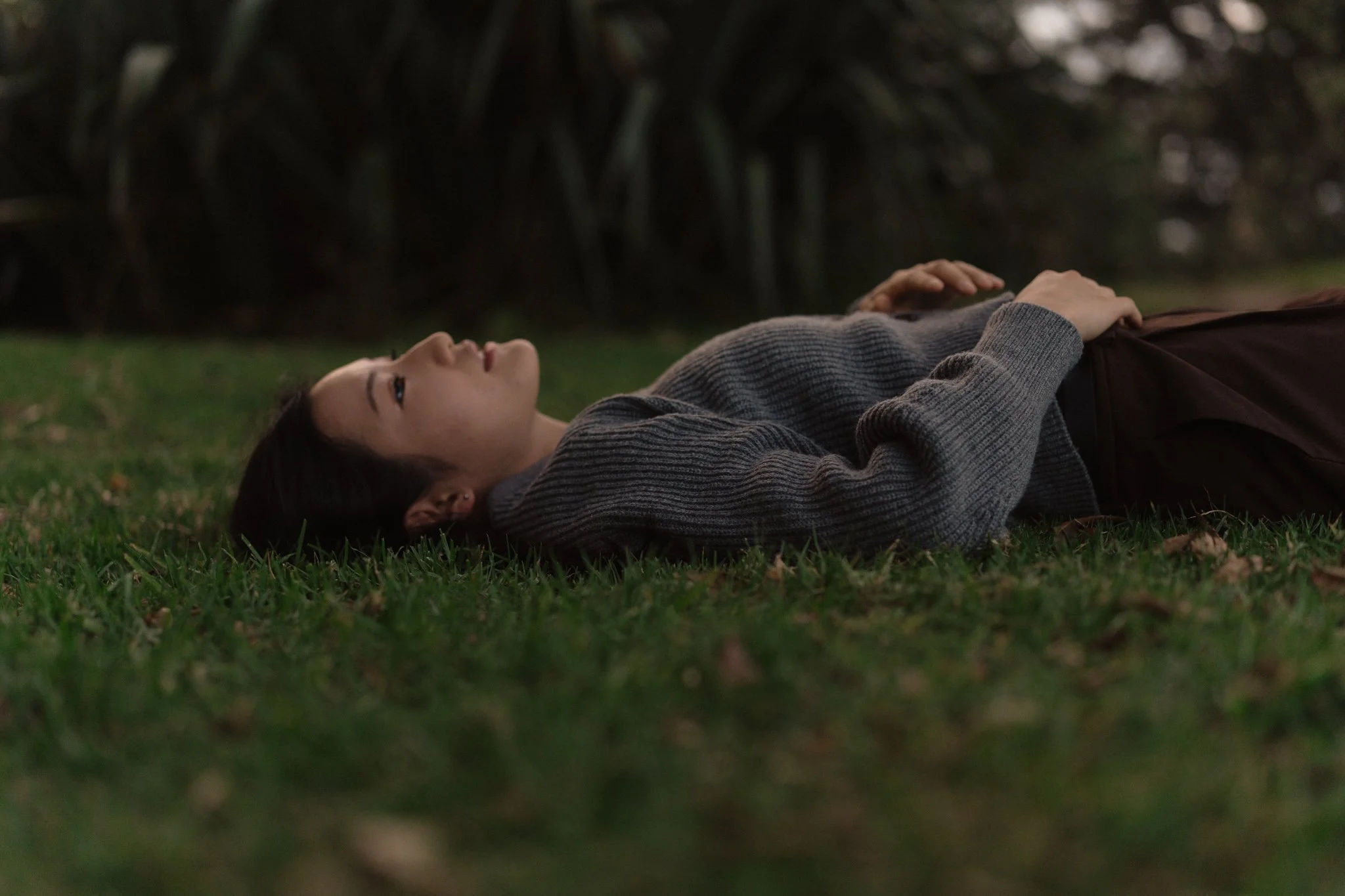 A woman lying on the grass outdoors, wearing a gray sweater and dark pants, with trees and plants in the background.