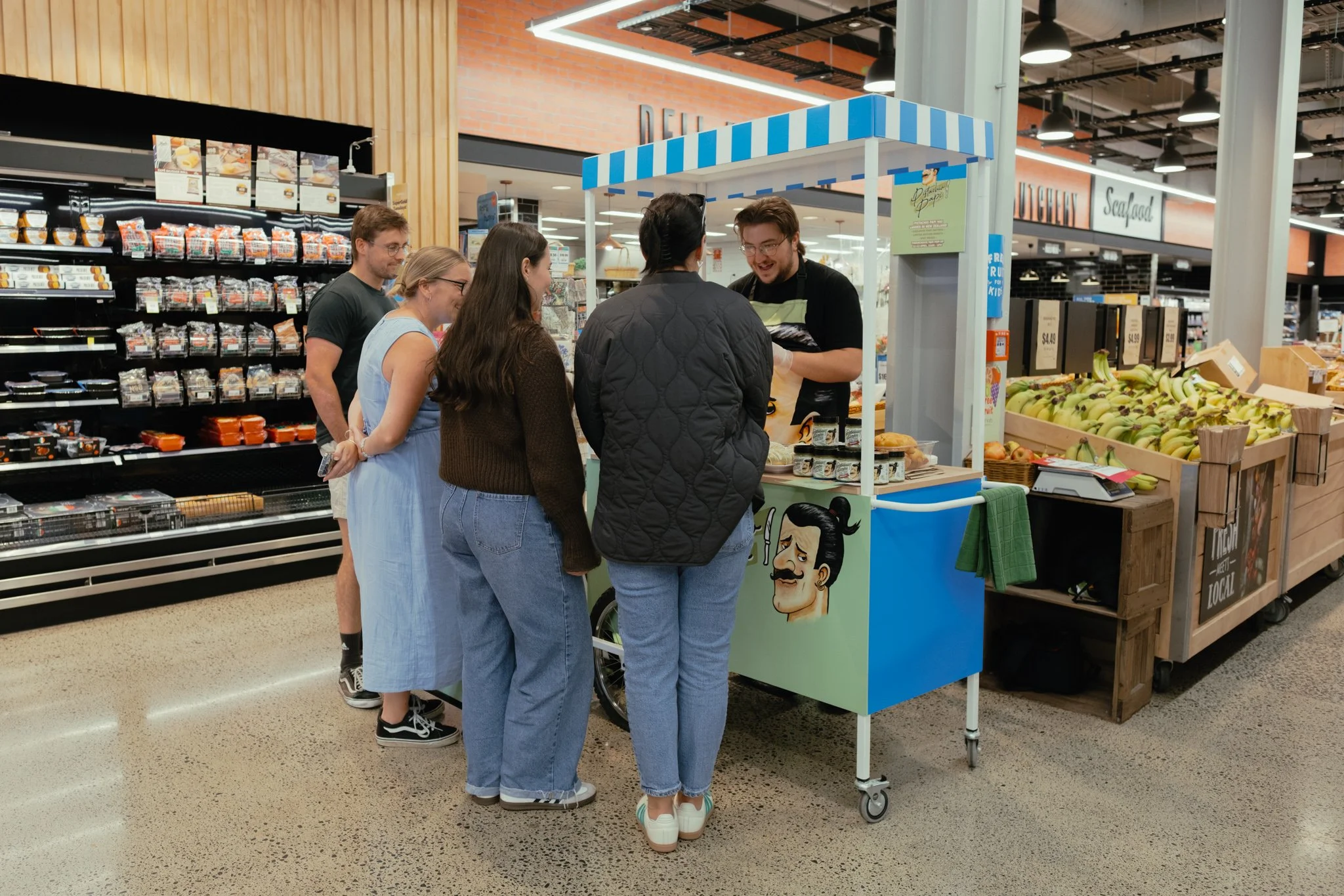 Group of five customers in line at a small sampling stall inside a grocery store.