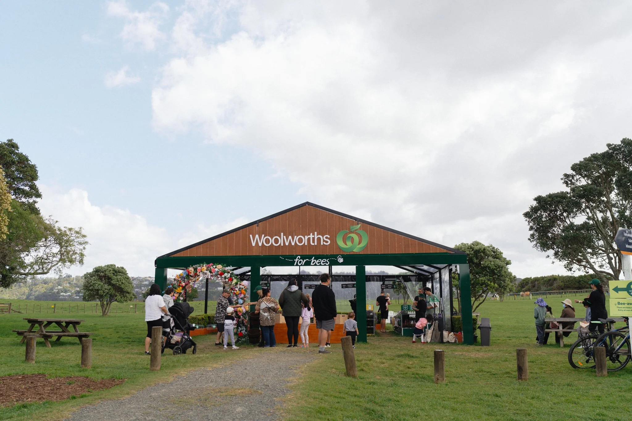 A crowd of people gather at a Woolworths bespoke supermarket for bees. People are sitting on benches, walking, and cycling nearby, with trees and an open grassy field in the background under partly cloudy skies.