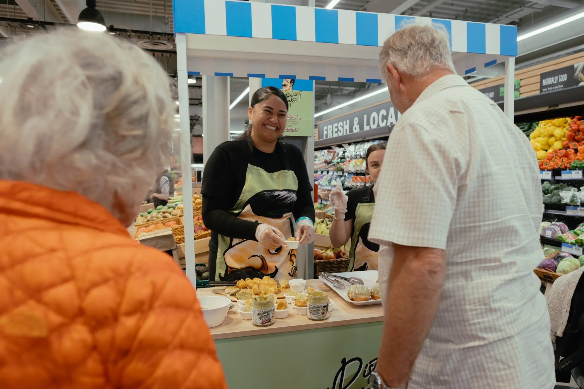 A woman at a sampling stand in a grocery store grins as she hands a sample to an older man in a yellow shirt.