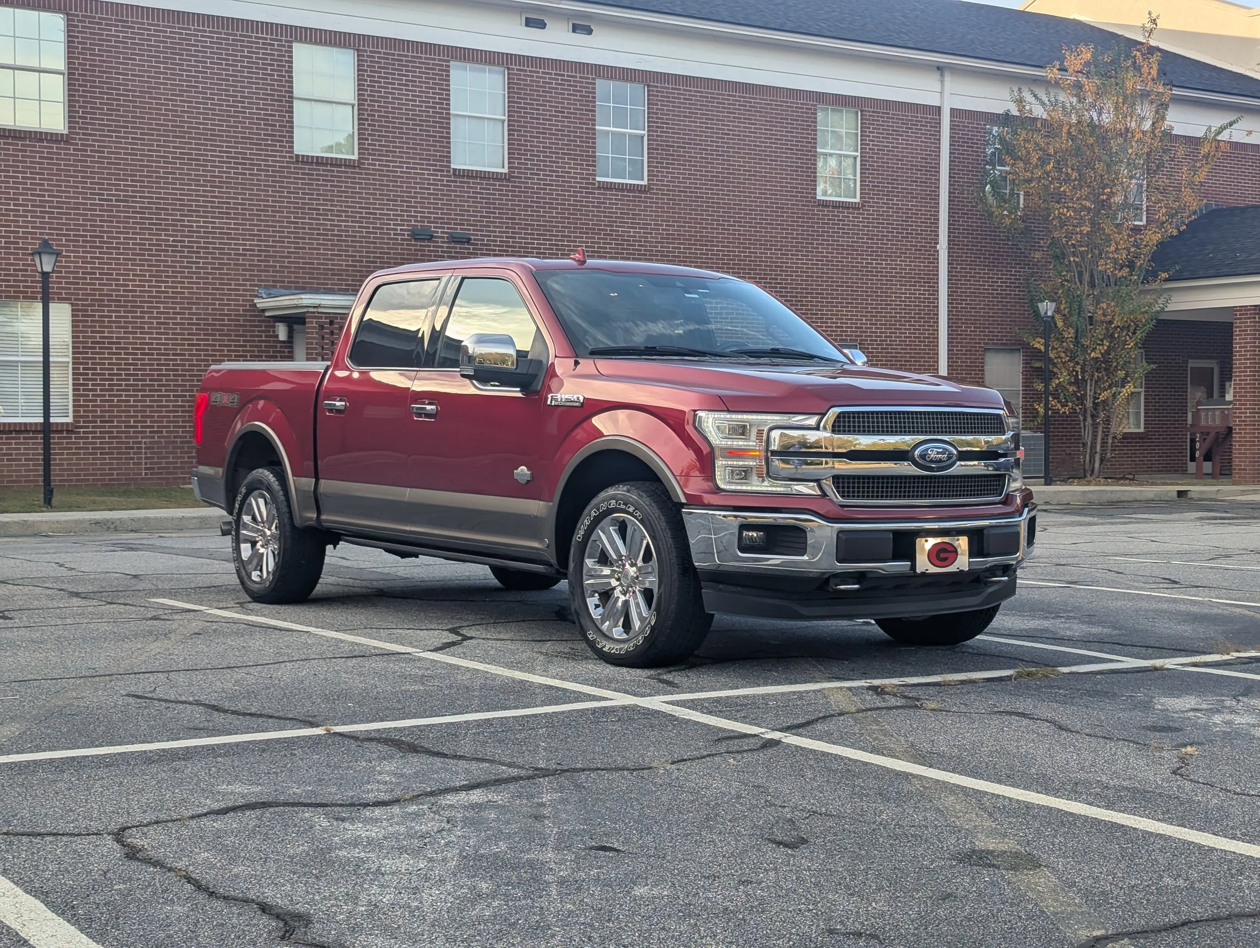 A red Ford pickup truck parked in an empty parking lot in front of a brick building with several windows and a small tree nearby.