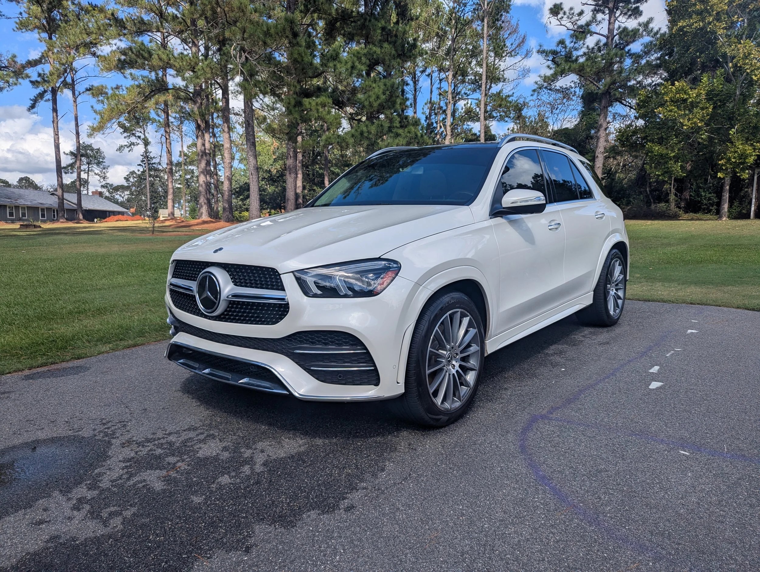 A white Mercedes-Benz SUV parked on a paved driveway in front of a lush grassy area with tall pine trees and a house with a gray roof in the background. The sky is partly cloudy with some blue visible.