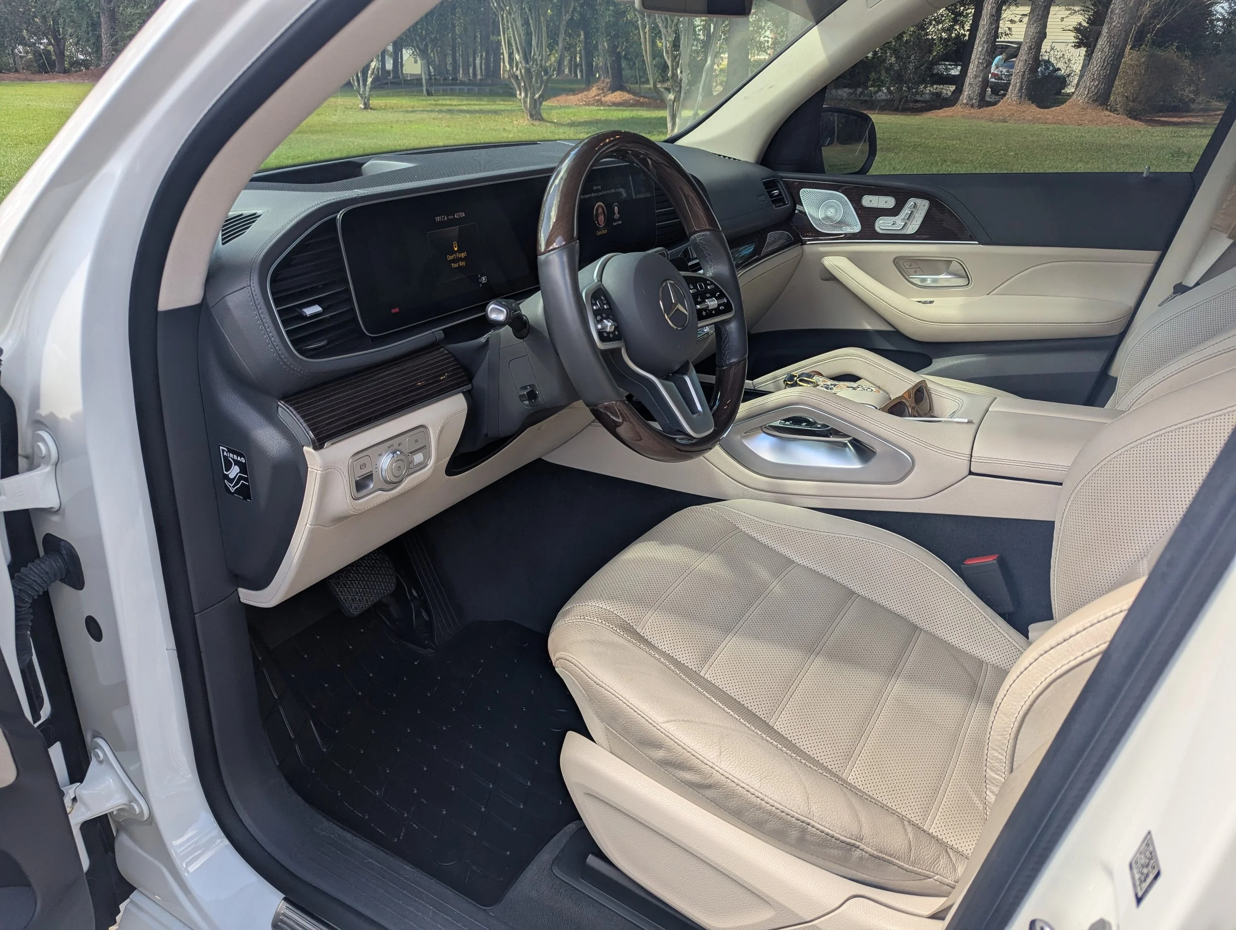 Interior of a luxurious Mercedes-Benz vehicle with beige leather seats, a sleek black dashboard, and a wood-trim steering wheel, parked outdoors with trees and a grassy area visible through the windshield.