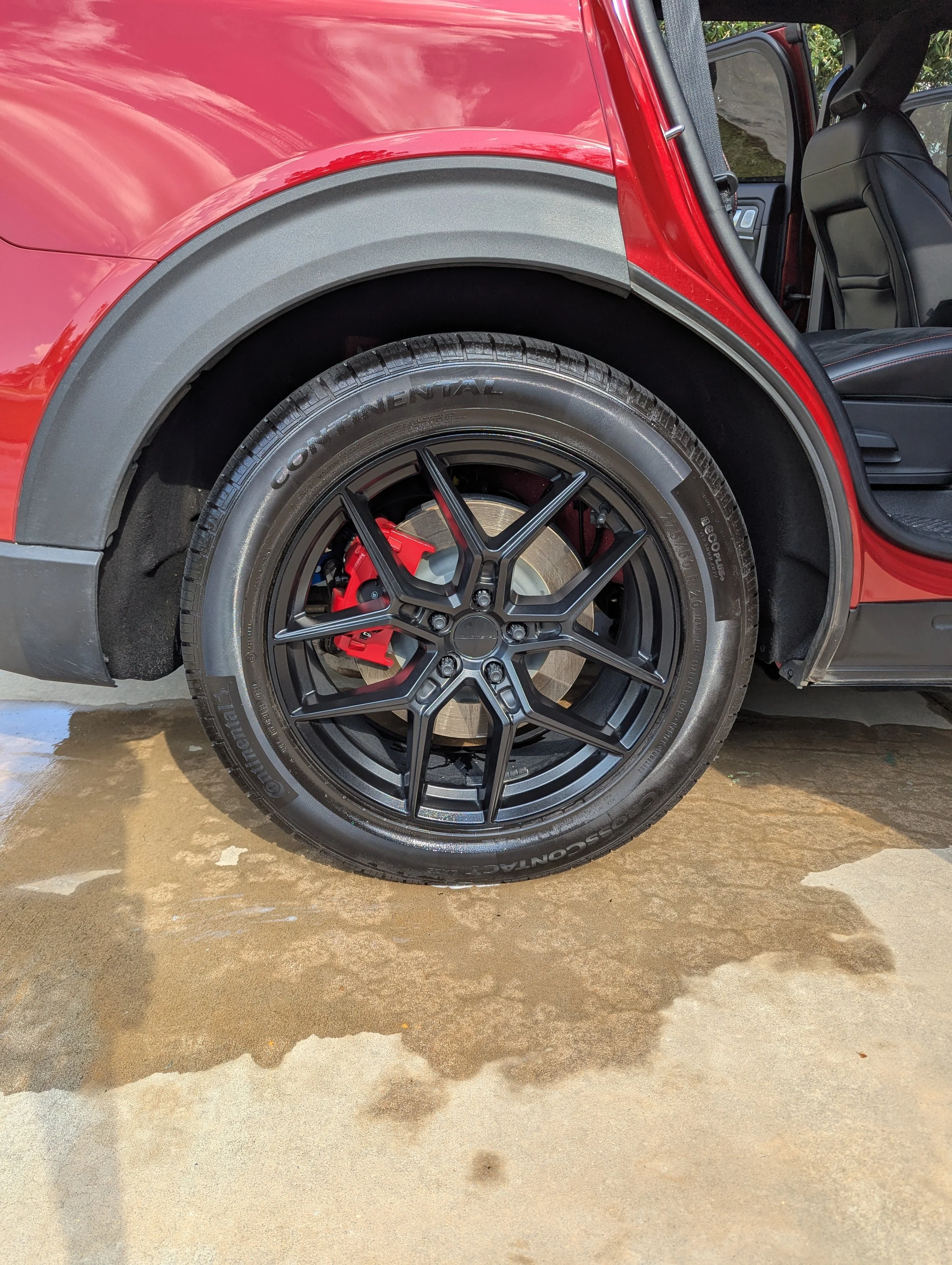 Close-up view of a red vehicle wheel with a low-profile tire, black alloy rim, and red brake caliper, on a wet concrete surface outside in daylight.