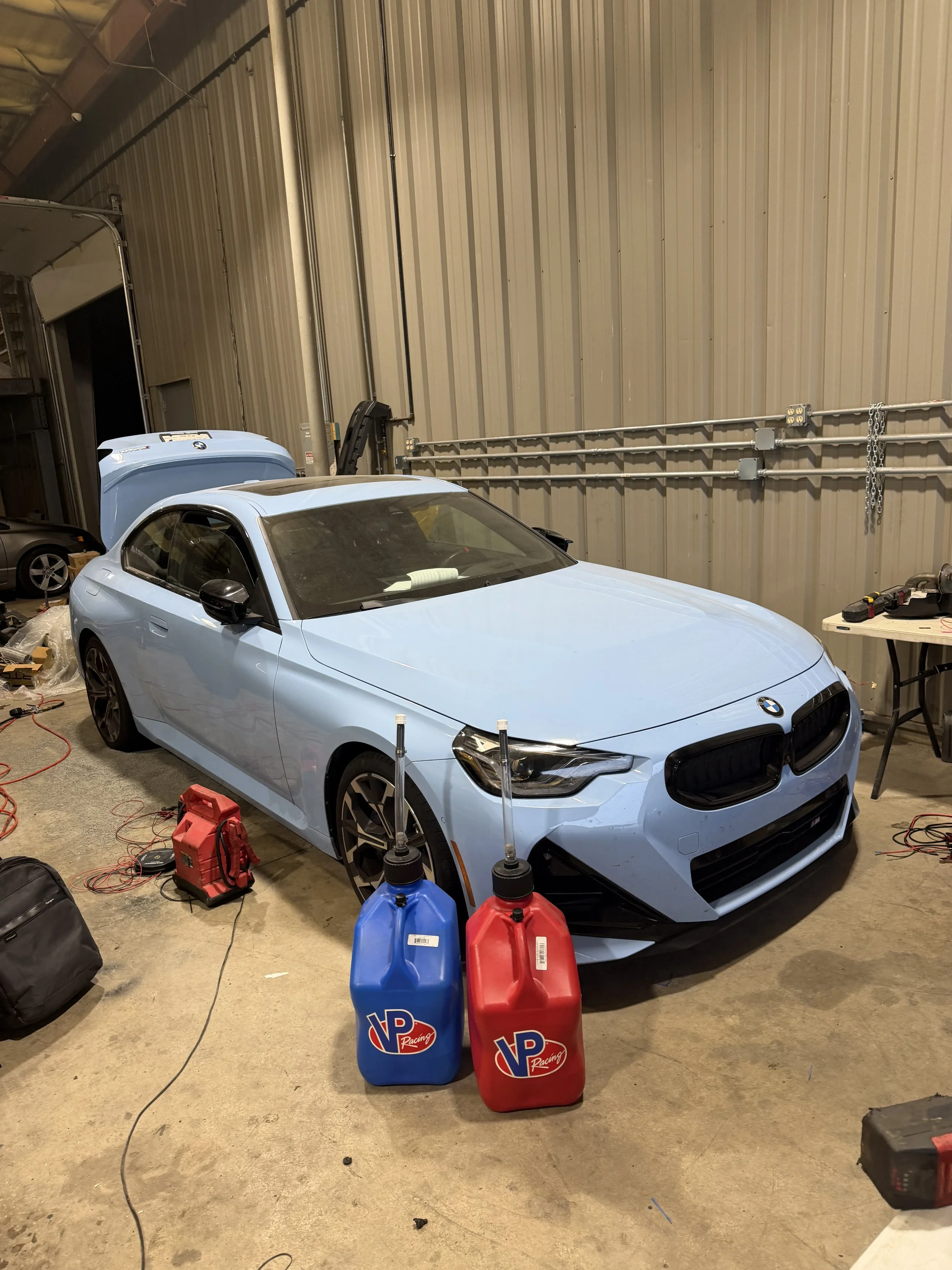 Light blue sports car in a garage with tools and supplies around, two large VP Racing fuel containers in front.