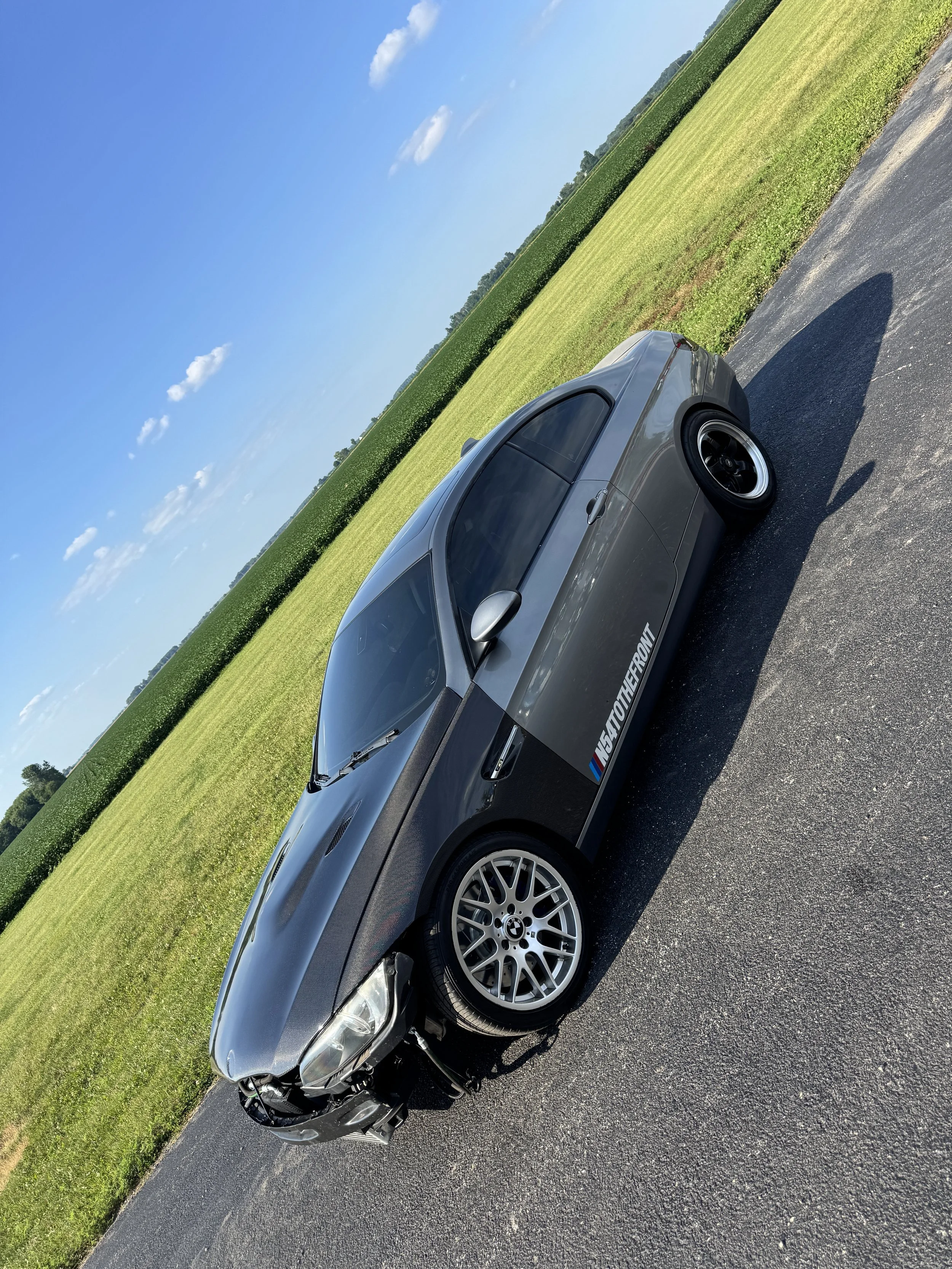 Silver BMW car with low profile tires parked on asphalt near grassy field with trees in the background under a blue sky with a few clouds.