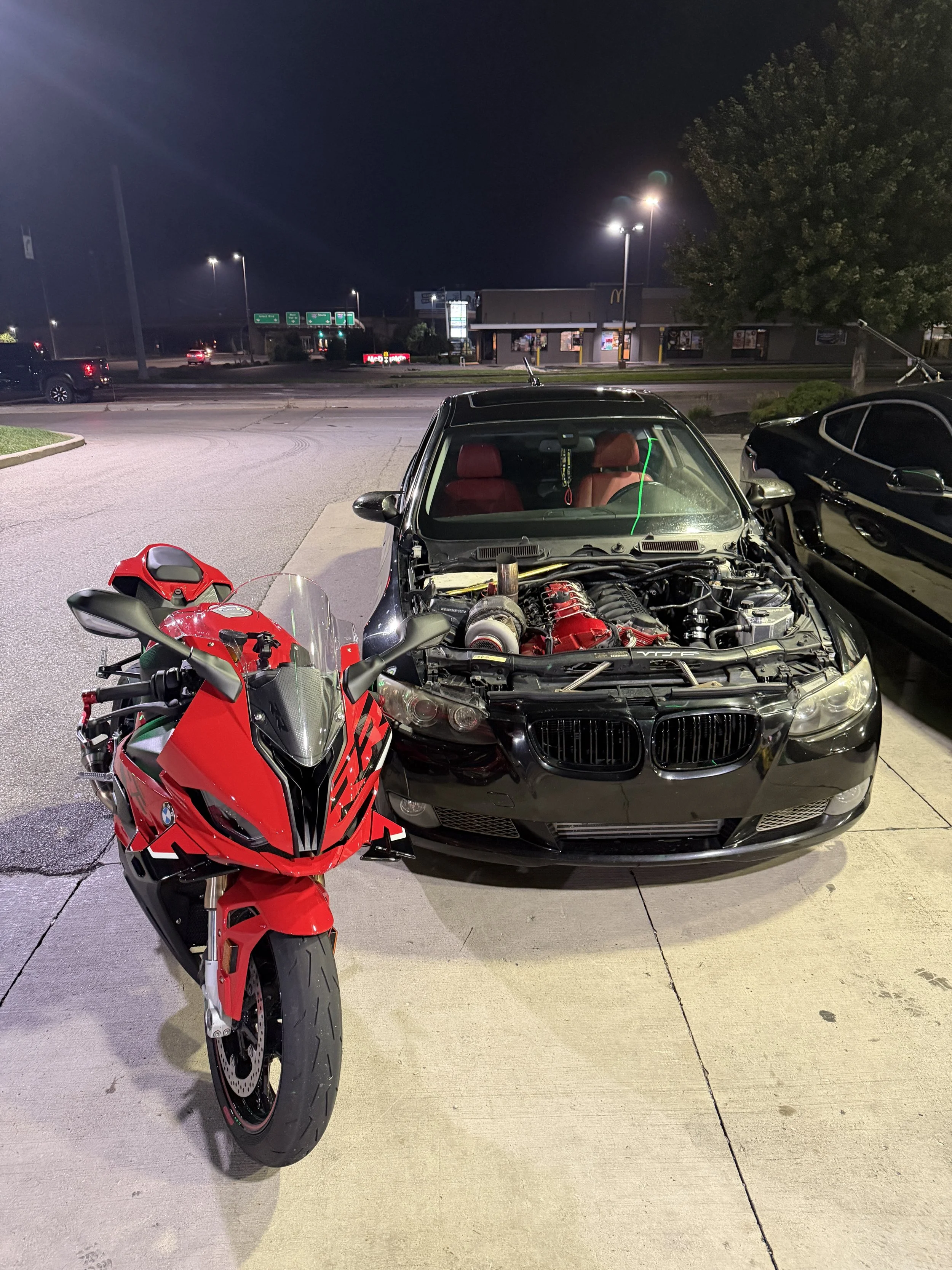 Nighttime parking lot scene with a red motorcycle in the foreground, and a black car with an open hood behind it. In the background, there is a strip mall with a McDonald's and other stores, illuminated by bright lights.