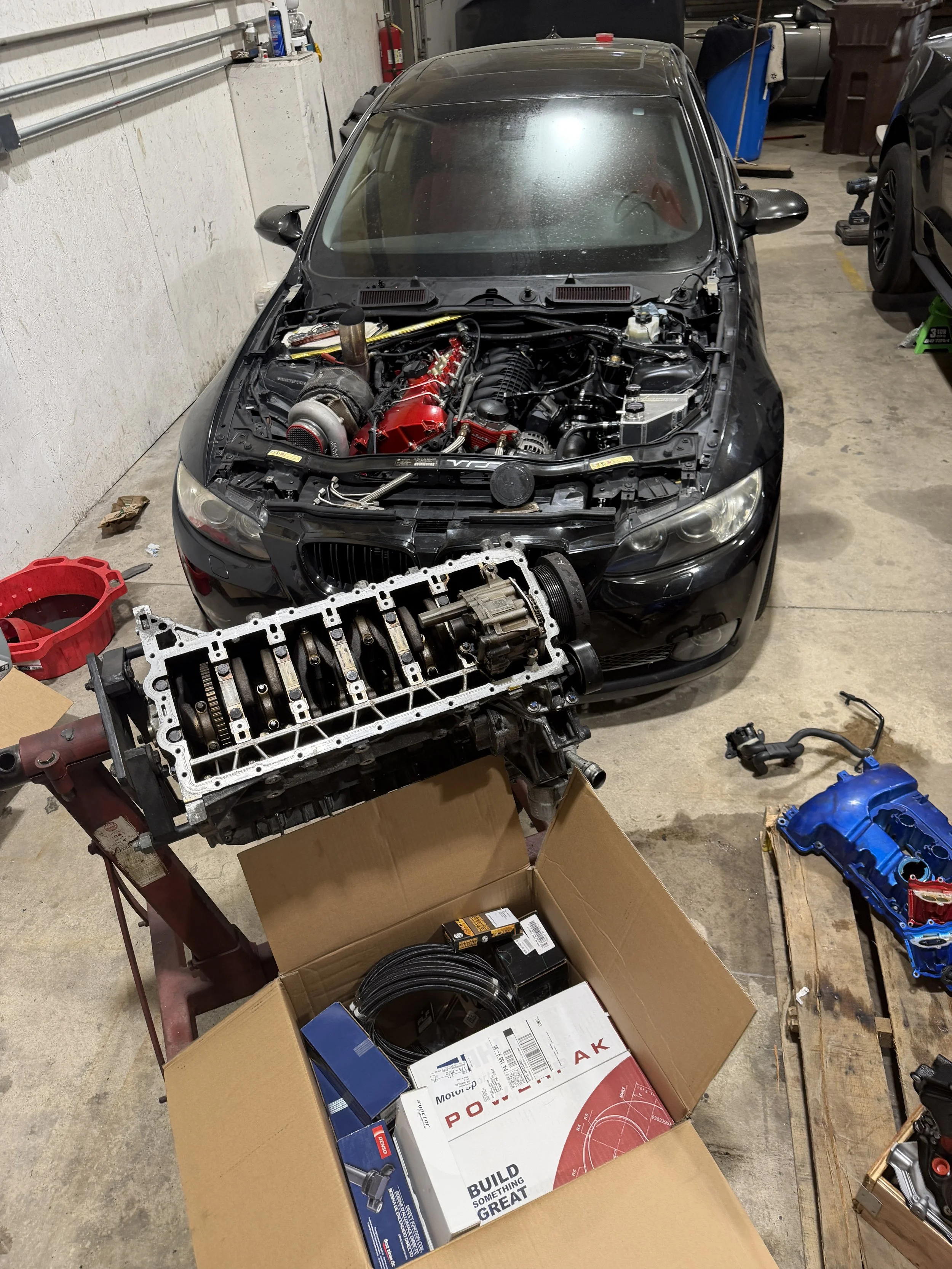 A black sports car with an open hood is undergoing engine work in a garage. The engine bay is partly disassembled, and various auto parts and tools are scattered around, including a turbocharger and a new engine component box.