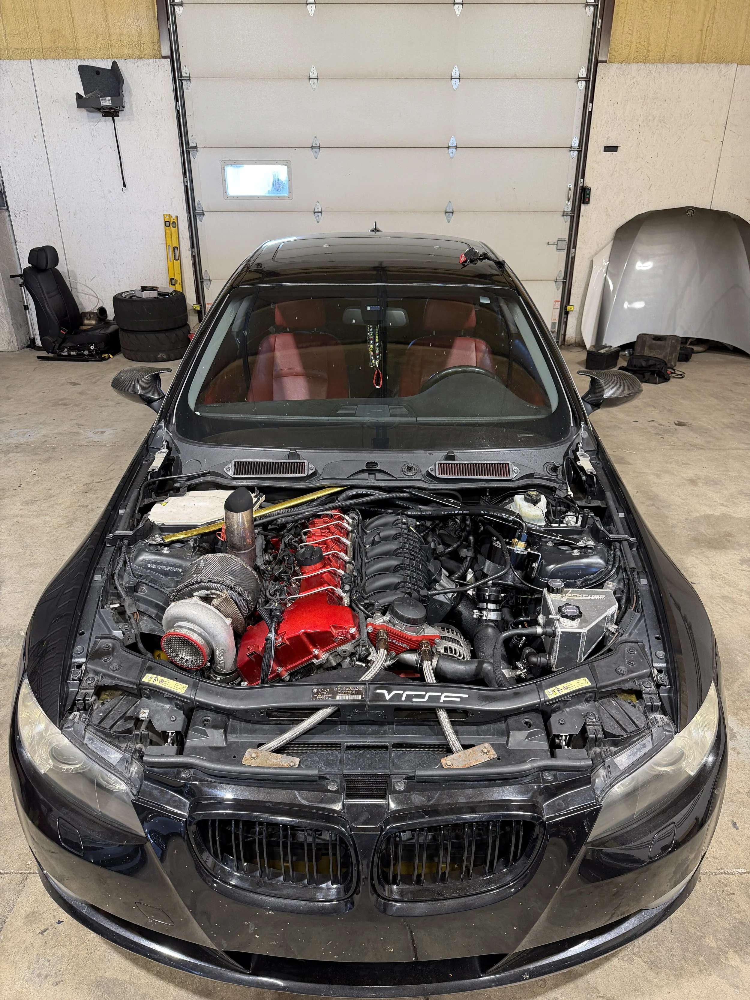 Front view of a black sports car with the hood open, exposing the engine, inside a garage.