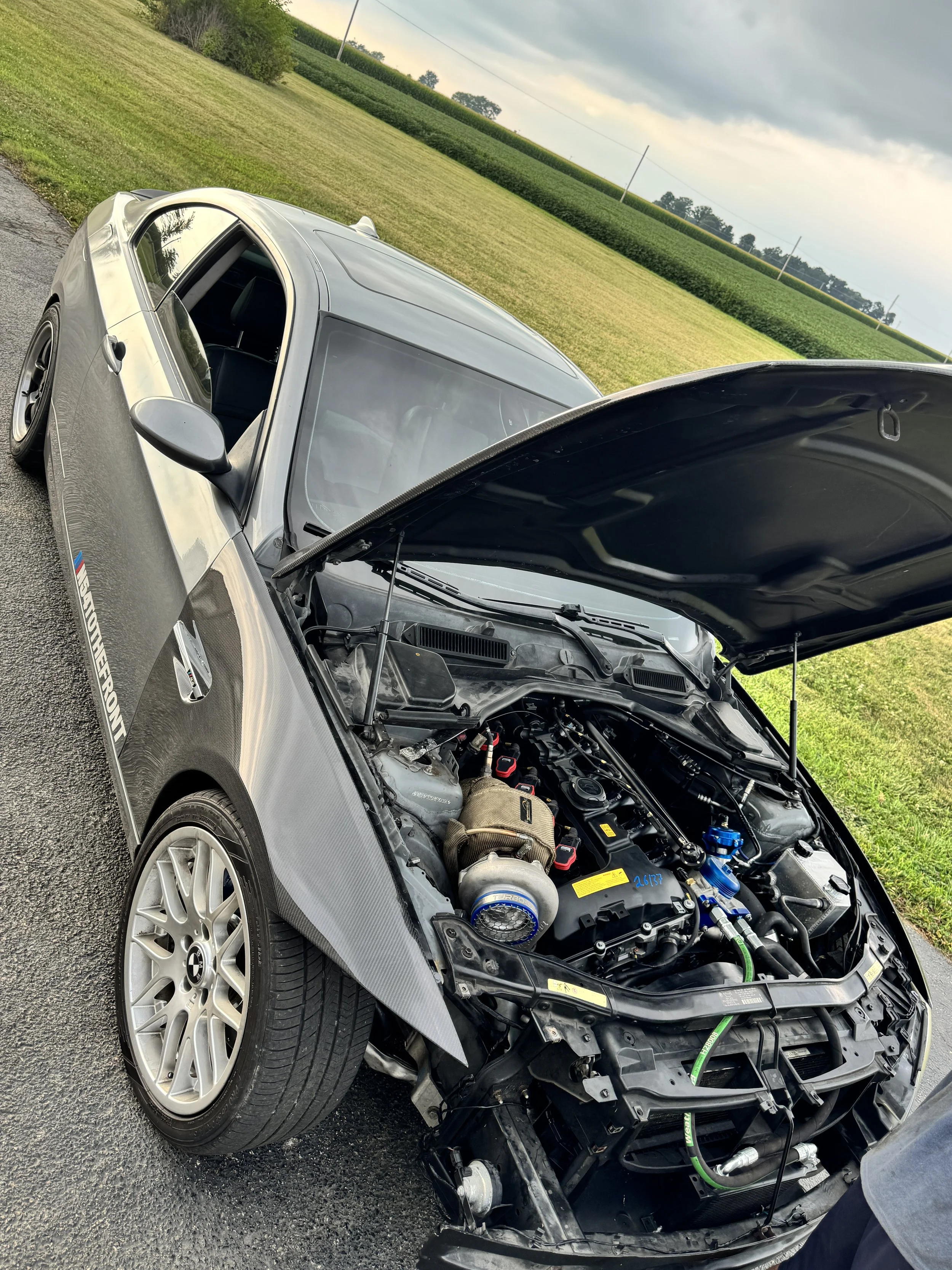 A silver sports car with its hood open, revealing a modified engine bay with aftermarket parts, parked on a paved surface near a grassy field under a cloudy sky.