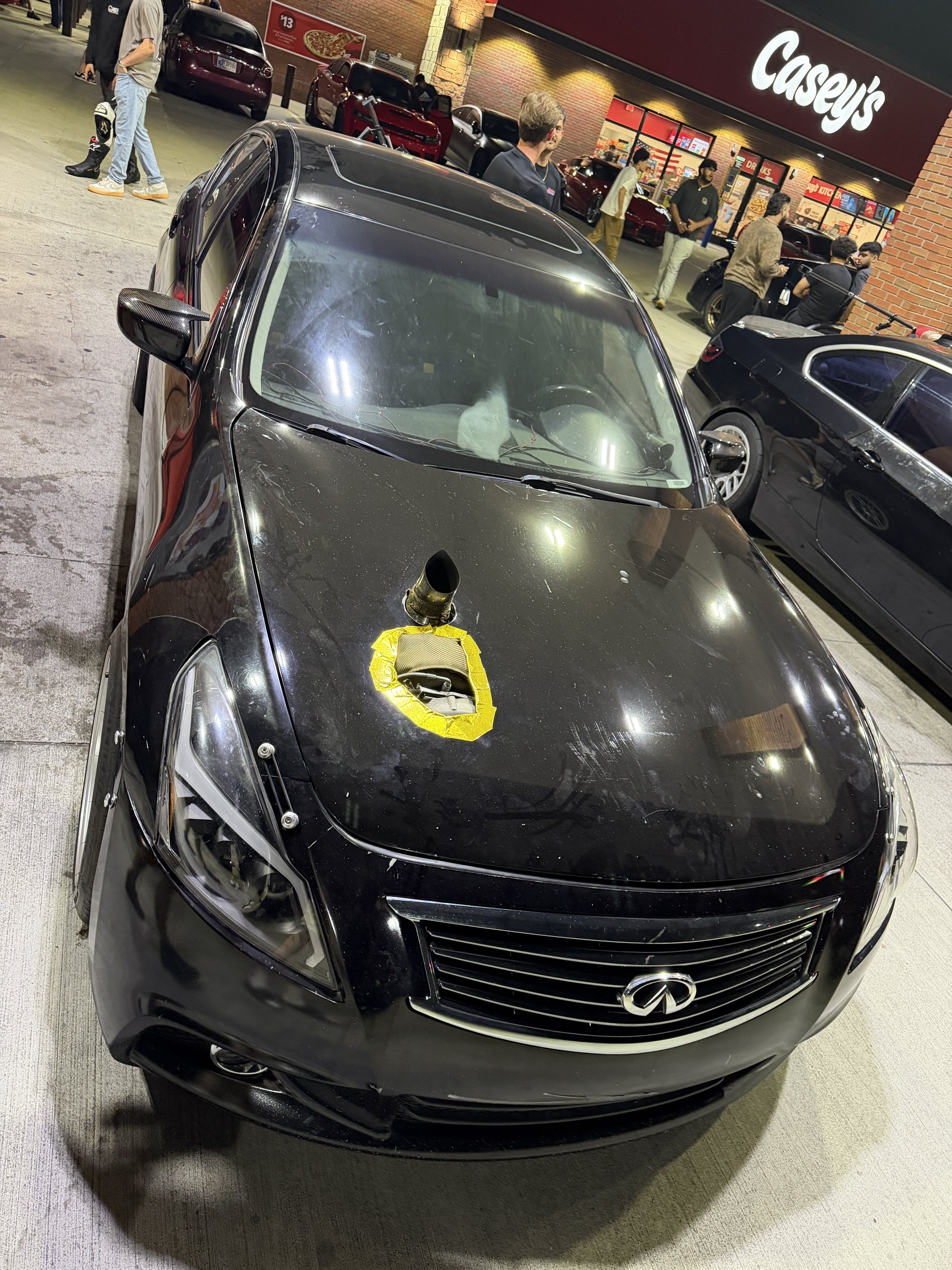 A black Infiniti car parked outside a Casey's store at night. The car has a yellow tape on the hood with a black object attached. Several people are gathered at the store entrance, and other cars are parked nearby.