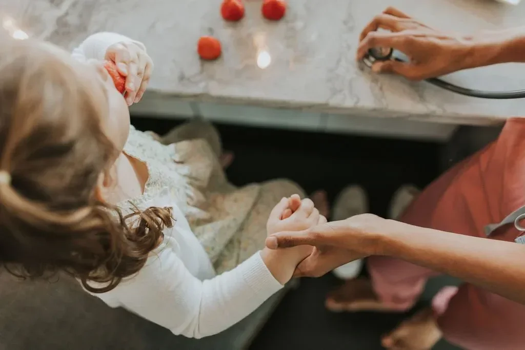 Overhead view of a parent holding a toddler’s hand at a kitchen table while the child eats a strawberry.