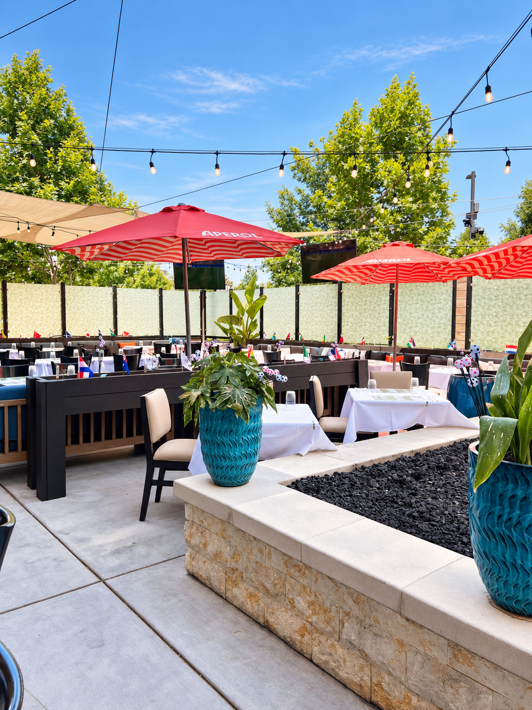 Outdoor restaurant patio with tables covered in white tablecloths, decorated with small flags. Red umbrellas and string lights are overhead. Potted plants and trees surround the area, creating a lively dining atmosphere.