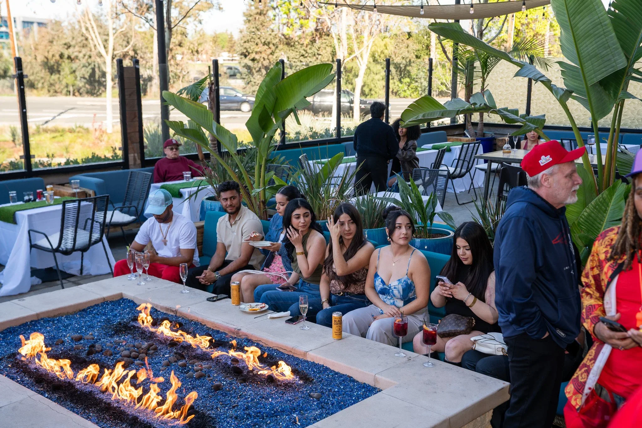 People dining at an outdoor restaurant with a patio fire pit, large tropical plants, and a view of the street in the background.