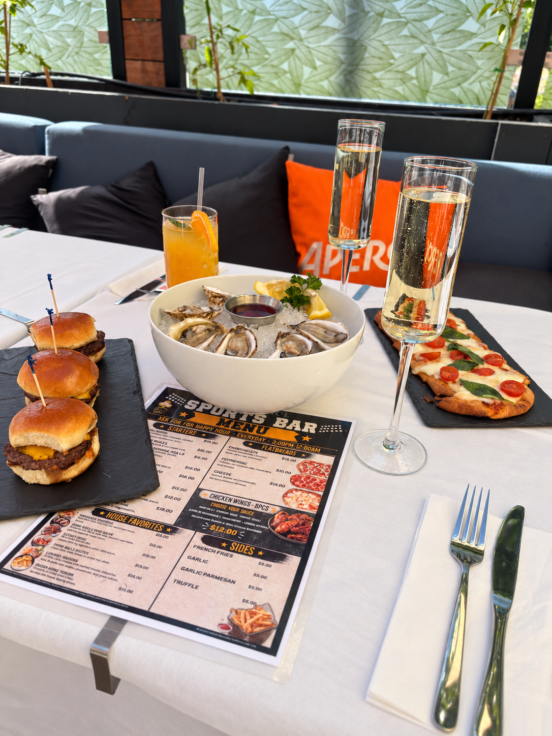 A table set with food and drinks including oysters on ice, sliders, a pizza, and beverages like champagne and orange juice, at a restaurant.