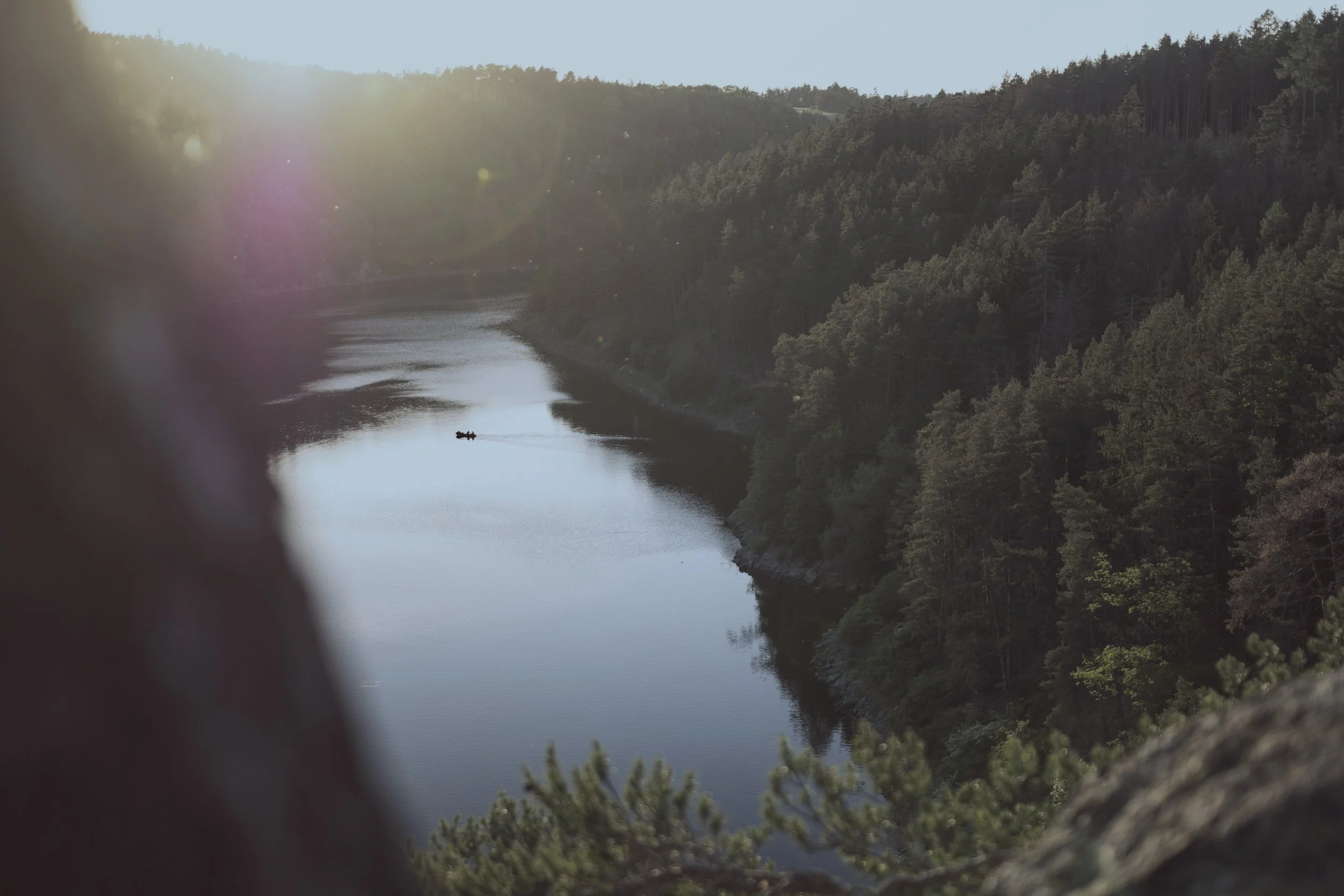 A river flowing through a forested landscape during the daytime, viewed through a opening with some shadows in the foreground.