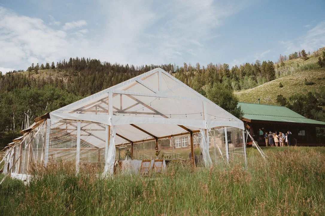 Outdoor wedding reception setup with a white tent and a separate green-roofed cabin, surrounded by grassy fields and wooded hills under a partly cloudy sky.