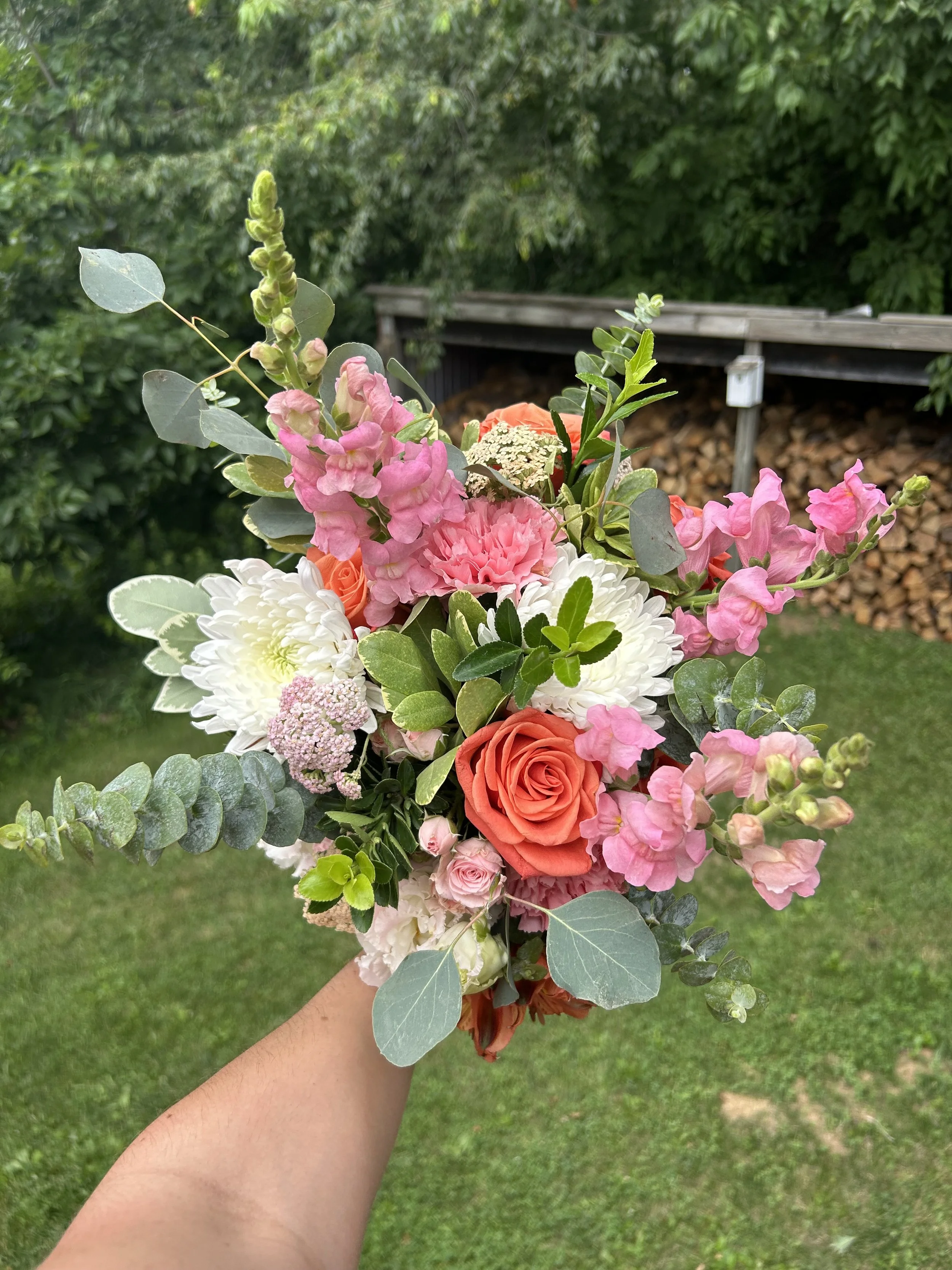 A bouquet of pink, orange, and white flowers held by a person's hand outdoors, with a green grassy area and a wooden structure in the background.