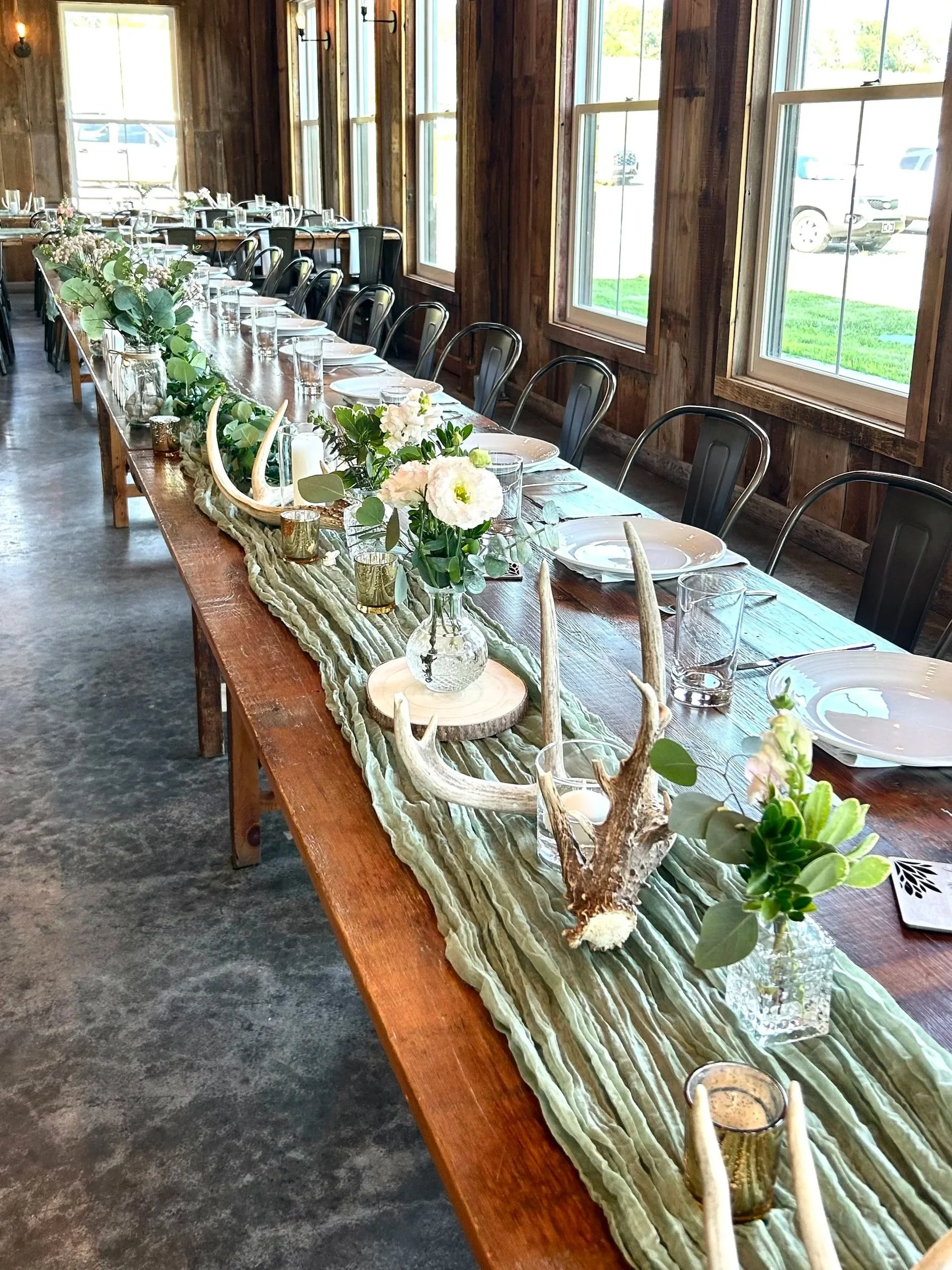 Long wooden table decorated with a green and beige table runner, glass candle holders, floral arrangements with white flowers and greenery, antler and wood accents, set with plates, glasses, and cutlery inside a rustic wooden room with large windows and natural lighting.