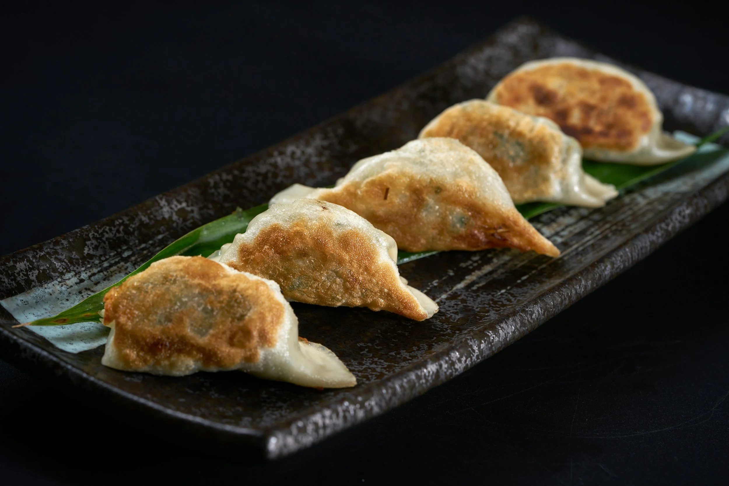 Close-up of several fried dumplings on a black rectangular plate with a green leaf underneath, set against a dark background.