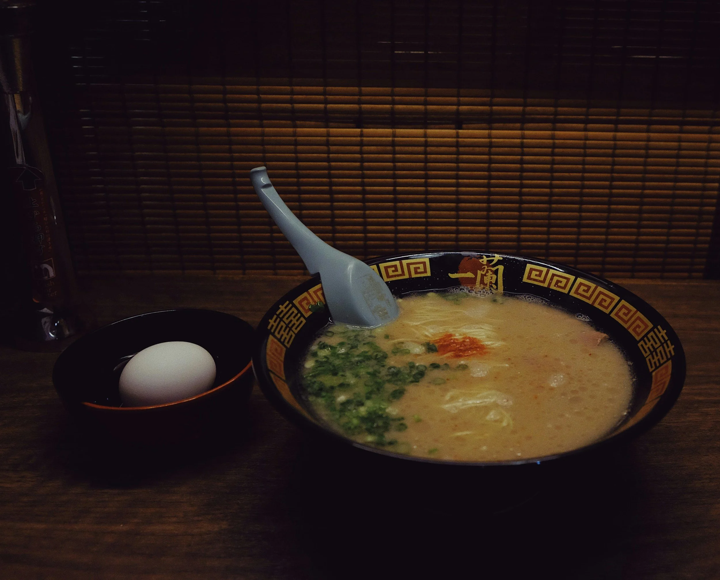 Bowl of ramen with green onions, egg, and broth on a wooden table, with a spoon, plate, and chopsticks nearby, against a bamboo backdrop.