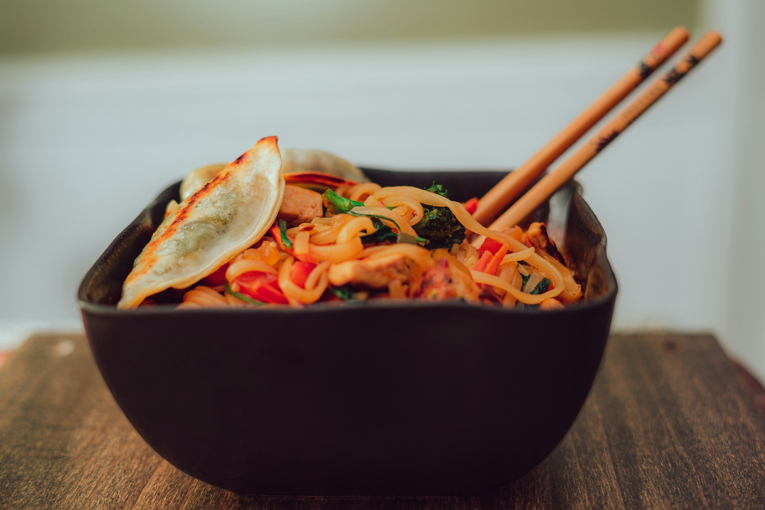 A black bowl filled with Asian-style stir-fried noodles, vegetables, and two grilled dumplings, with a pair of chopsticks resting on the side, on a wooden surface.