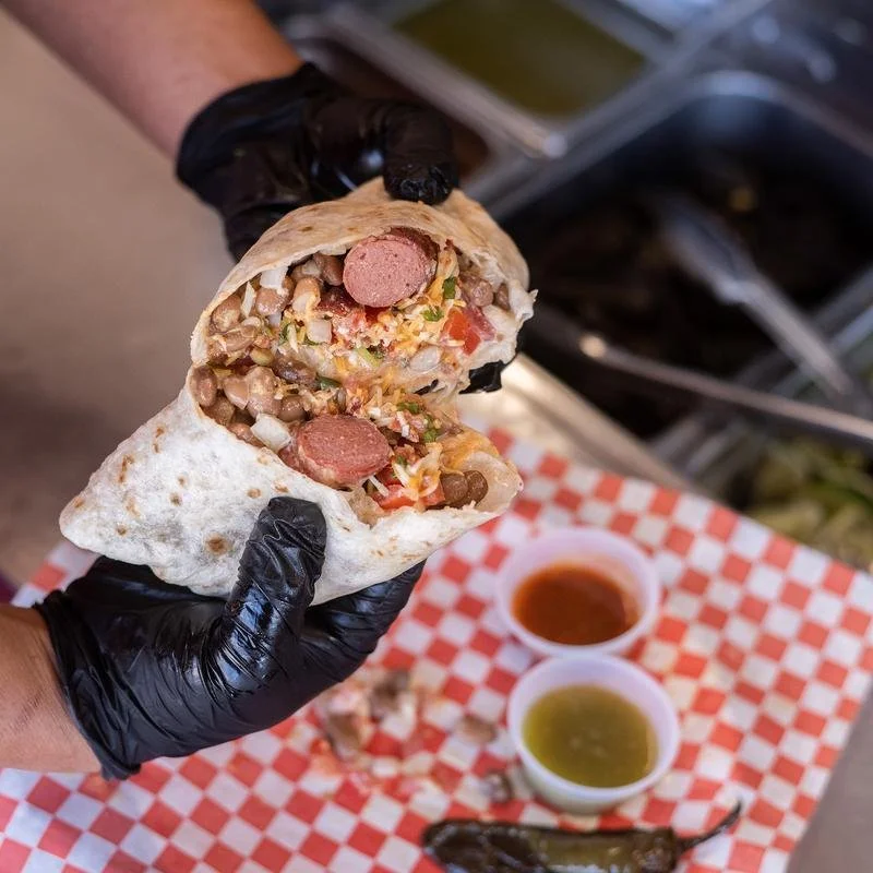 Person wearing black gloves holding a sliced burrito filled with sausage, beans, lettuce, tomato, and other ingredients. The background includes two small bowls of sauce and a checkered paper-lined tray.