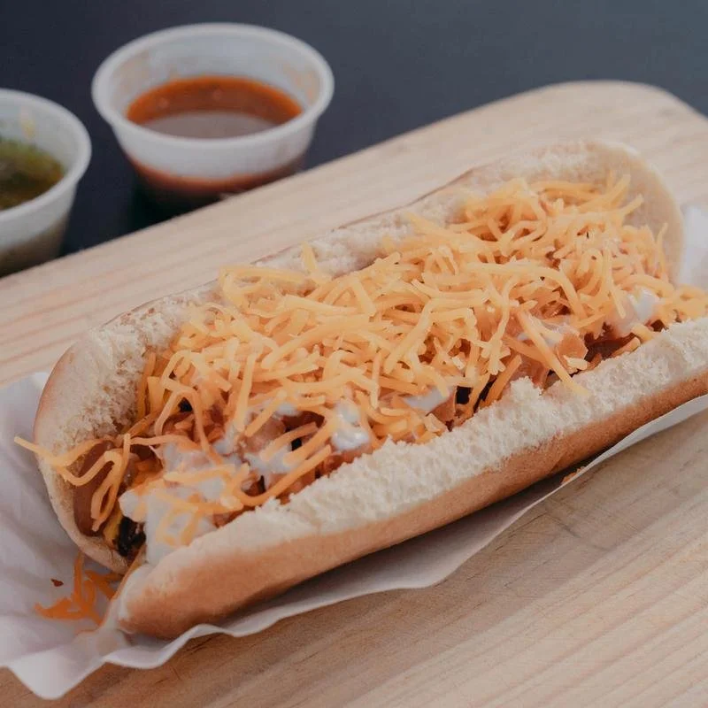 Philly cheesesteak sandwich with shredded cheese, on a hoagie bun, on a wooden serving board with two dipping sauces in the background.