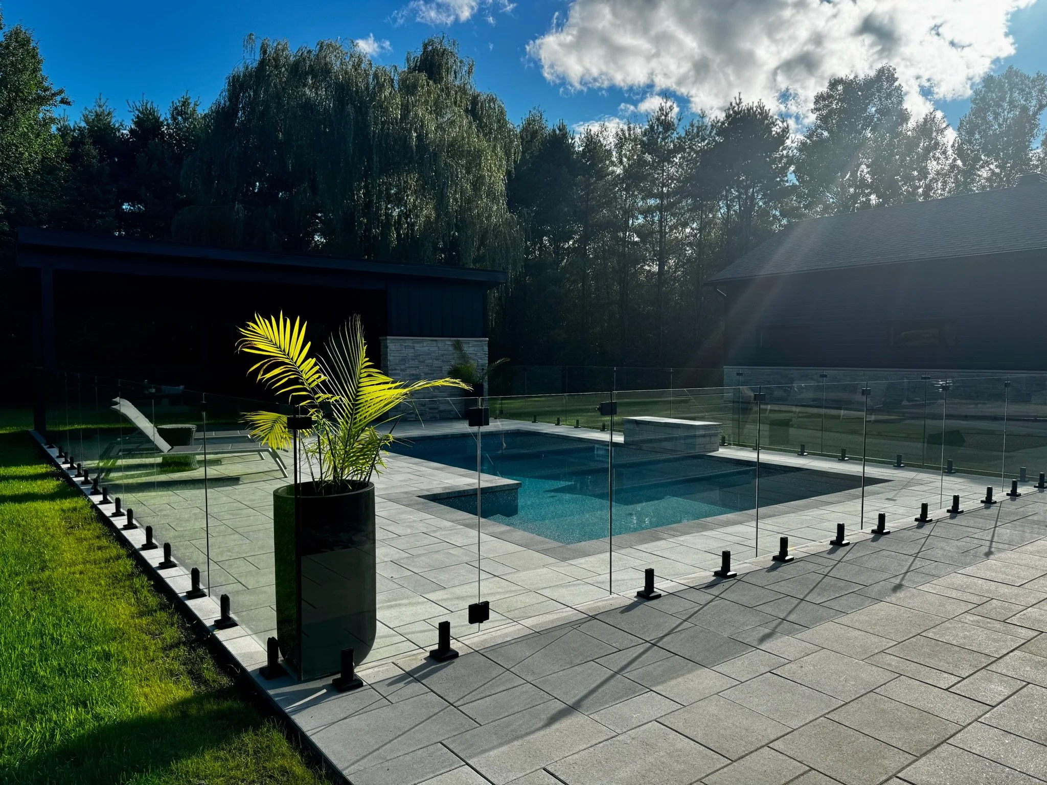 Piscine extérieure avec terrasse en pavés, entourée d'une clôture en verre, avec un grand pot de plantes vertes en premier plan. Dans le fond, une maison avec un toit en tuile et un ciel partiellement nuageux.