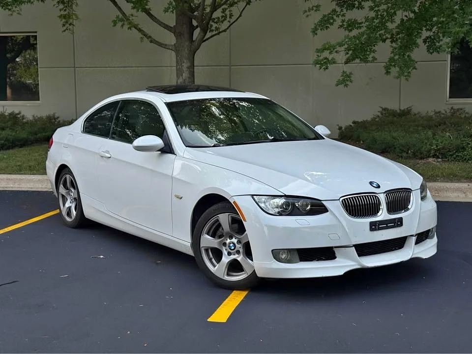 White BMW coupe parked in a parking lot next to a tree and bushes.