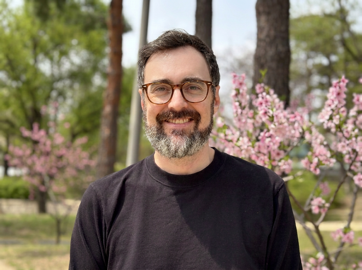 A smiling man with glasses and a beard outdoors, with pink flowering trees and green trees in the background.