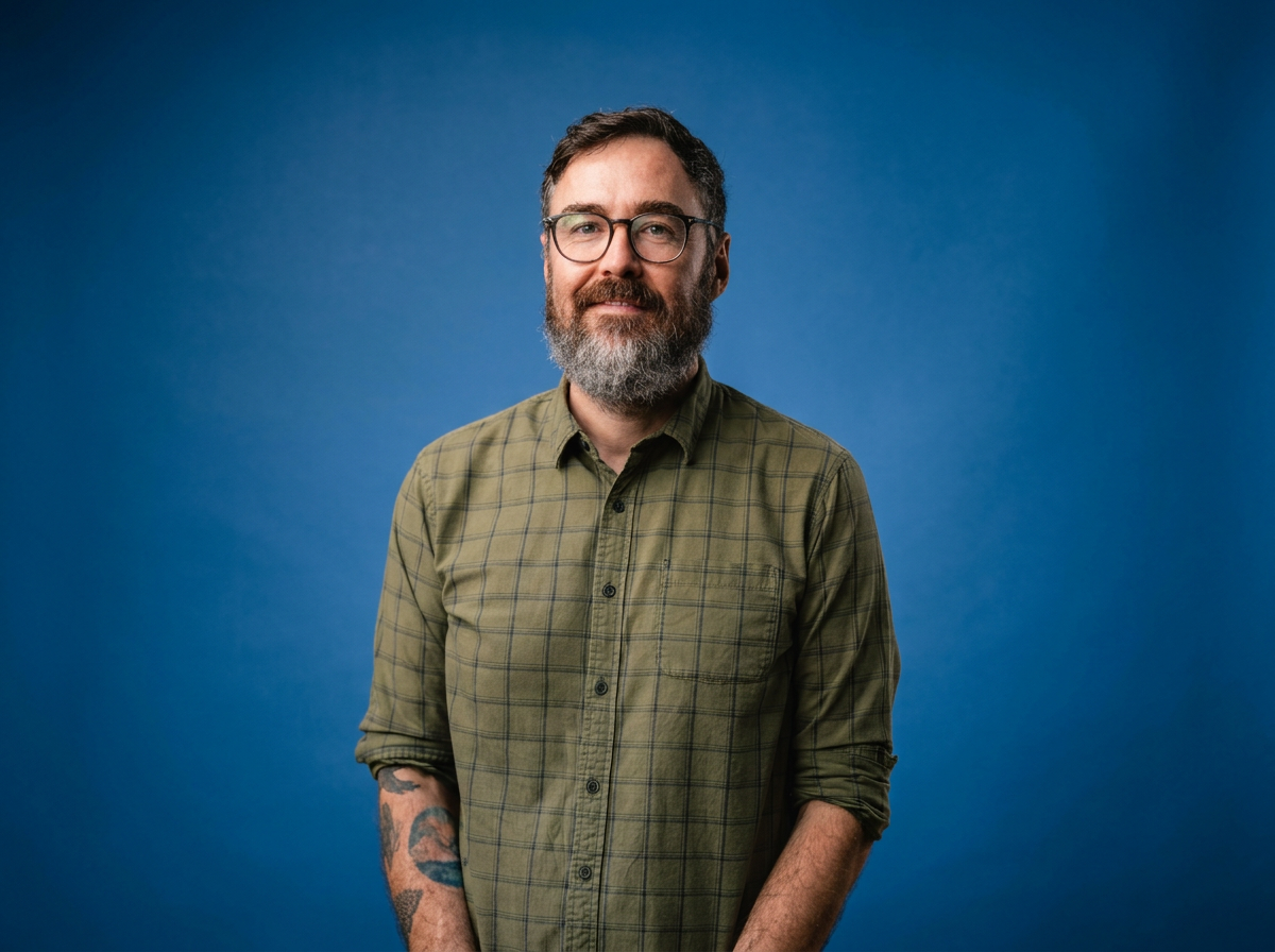 A man with glasses and a beard, wearing a green plaid shirt, standing against a blue background.