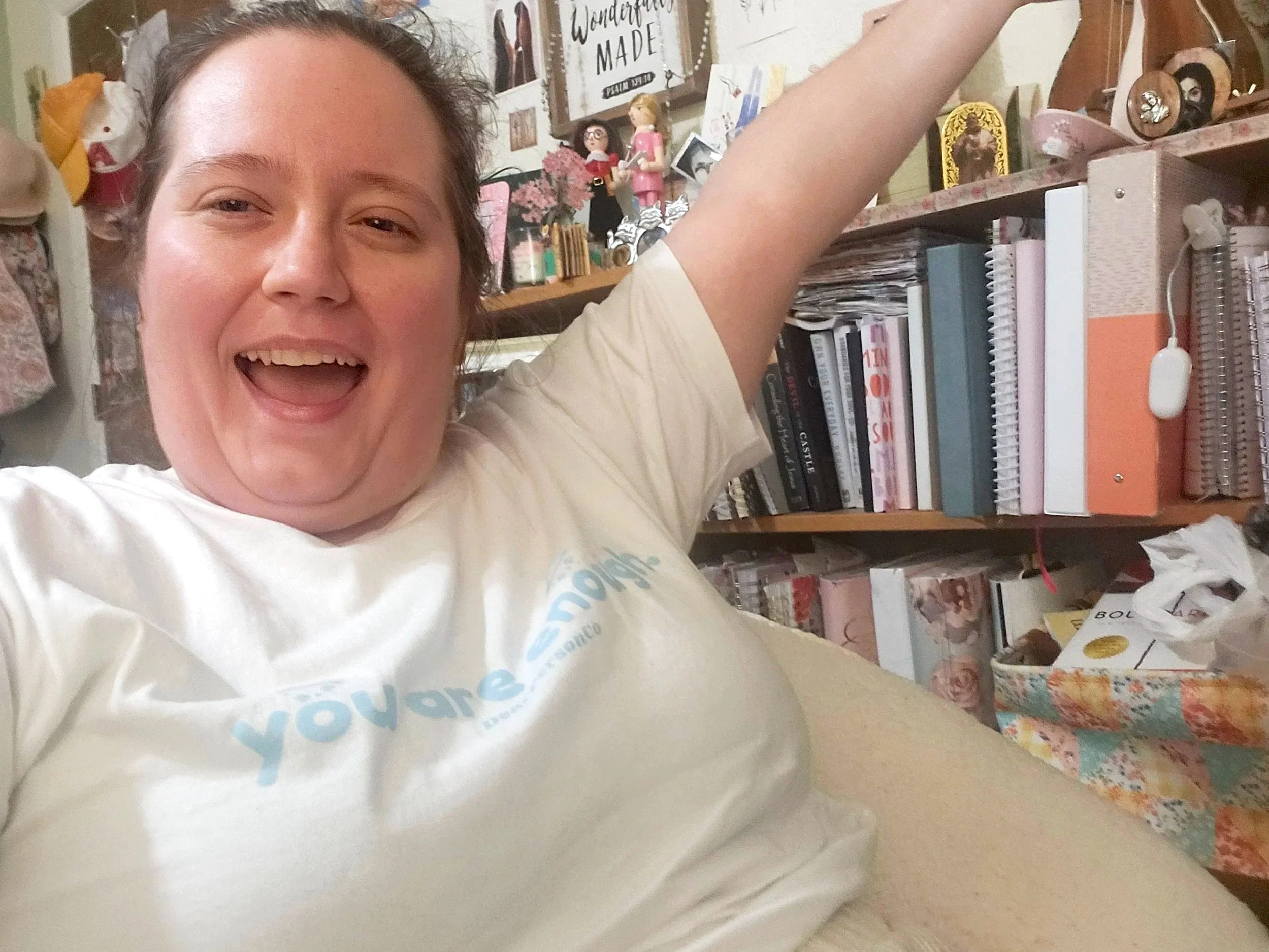 Close-up of a woman smiling and taking a selfie in front of a cluttered bookshelf filled with books, decorative items, and framed pictures.