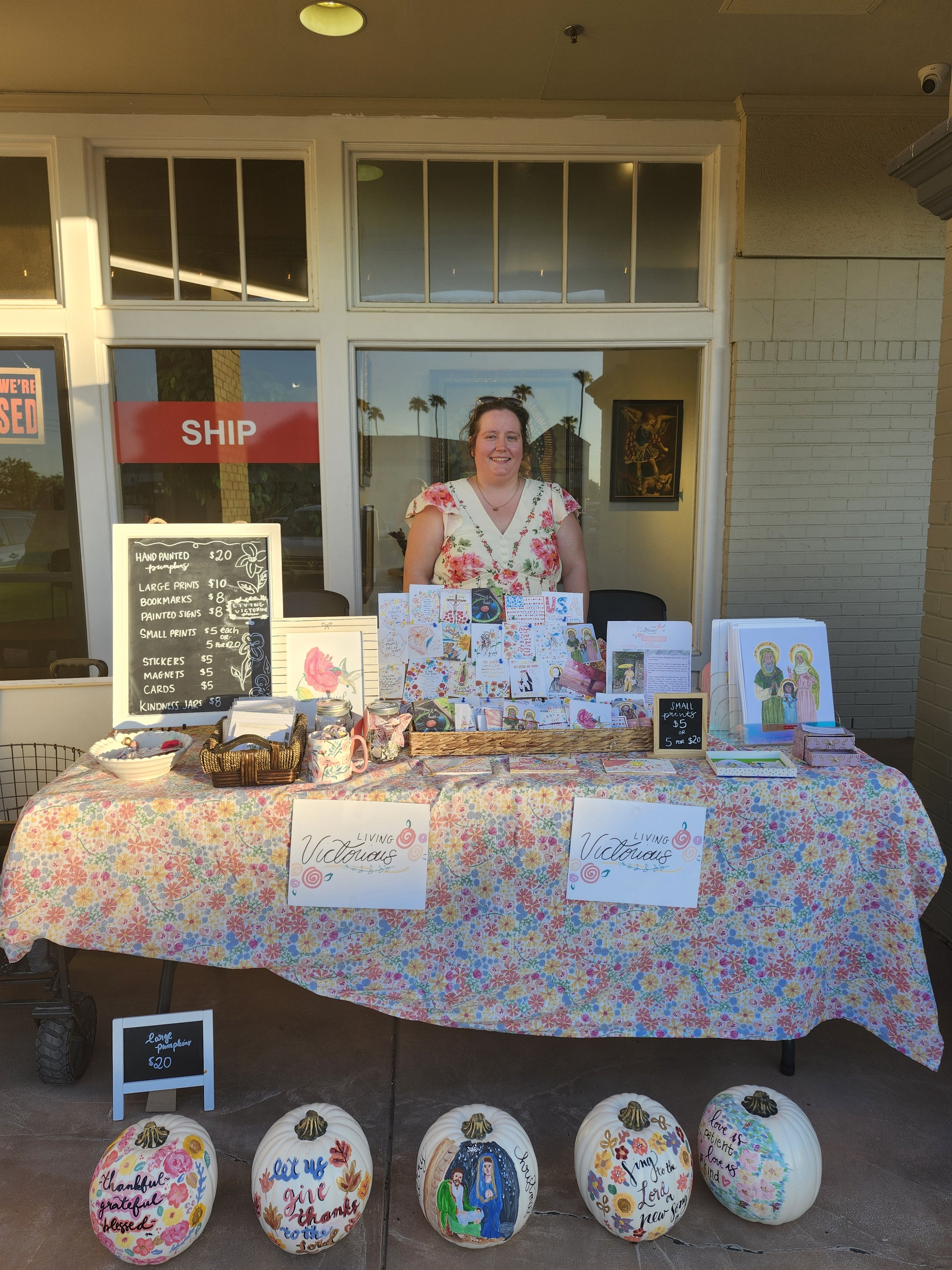 A woman standing behind a table with various handmade crafts, including cards and painted pumpkins, outside a storefront with large glass windows. The table is covered with a colorful floral tablecloth, and there are signs displaying prices for the items. Decorative pumpkins with painted artwork and messages are at the front of the table.