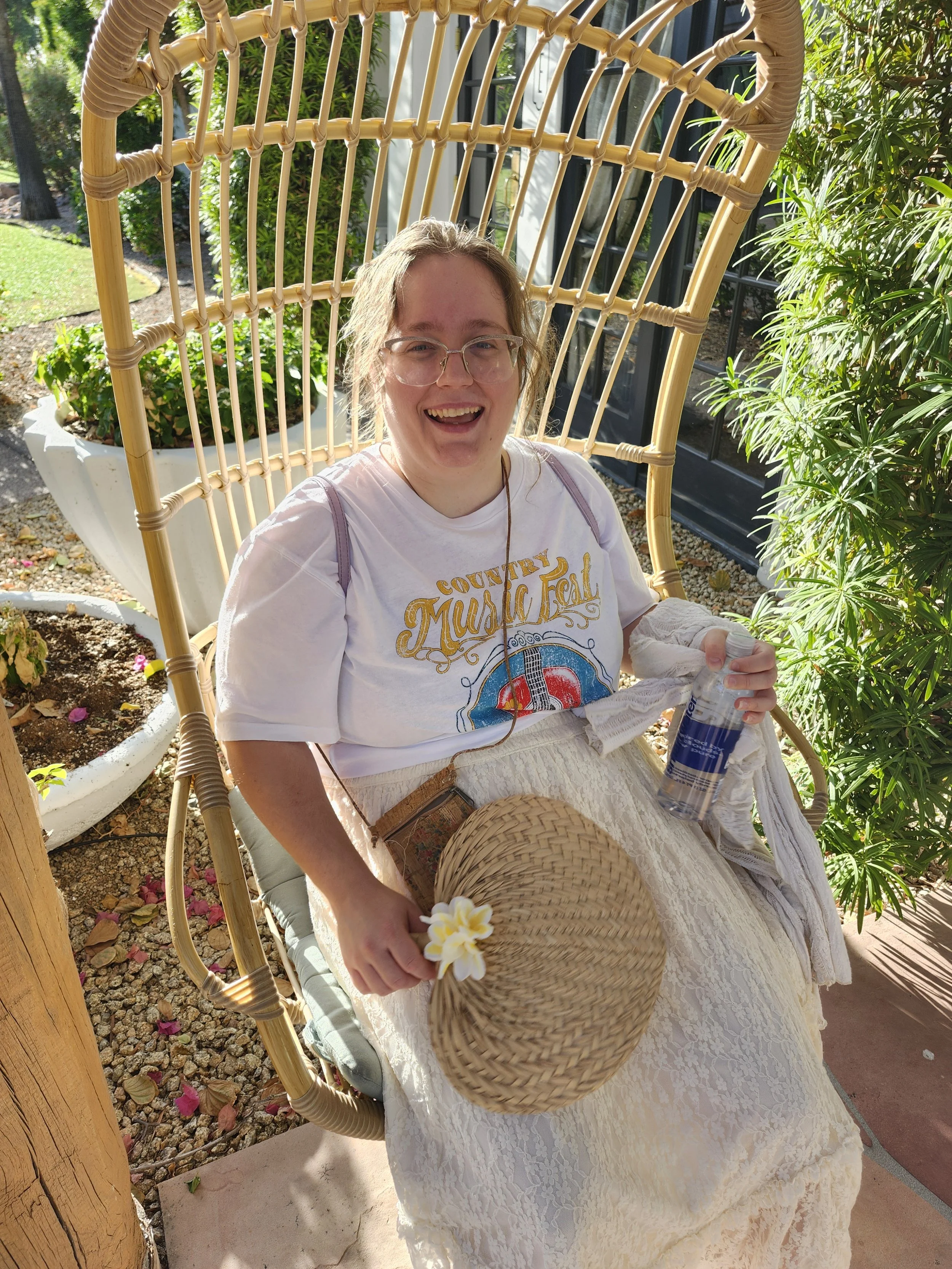 A smiling woman with glasses sitting on a wicker chair outdoors, holding a straw hat with a flower, with plants and a black door in the background.